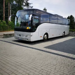 Srebrny autokar Mercedes-Benz na parkingu z kostki brukowej, widoczny znak ostrzegawczy dla dzieci w oknie.