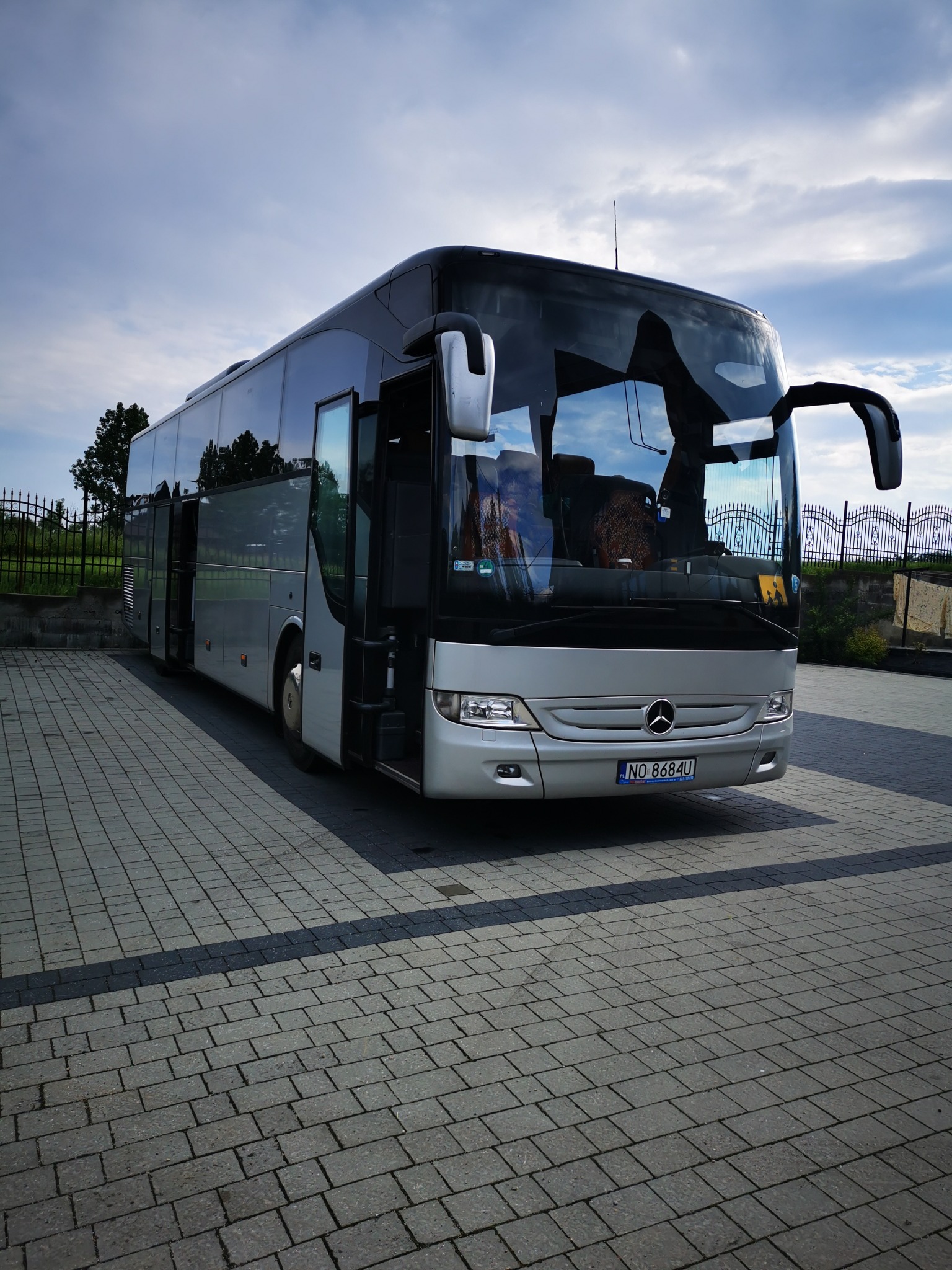 Srebrny autokar Mercedes z otwartymi drzwiami, zaparkowany na kostce brukowej, widok z przodu na tle błękitnego nieba z chmurami.