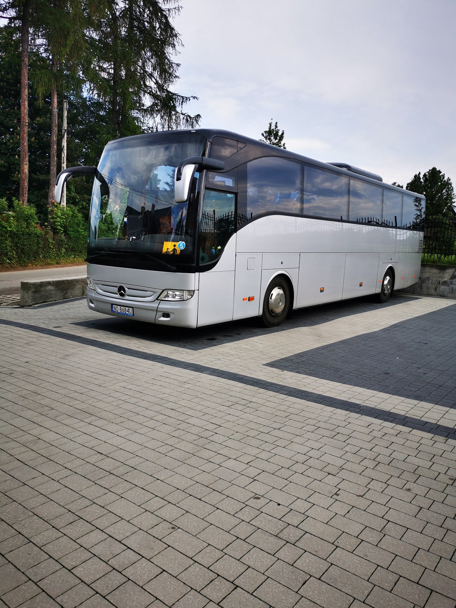 Srebrny autokar Mercedes-Benz na parkingu z kostki brukowej, widoczny znak ostrzegawczy dla dzieci w oknie.