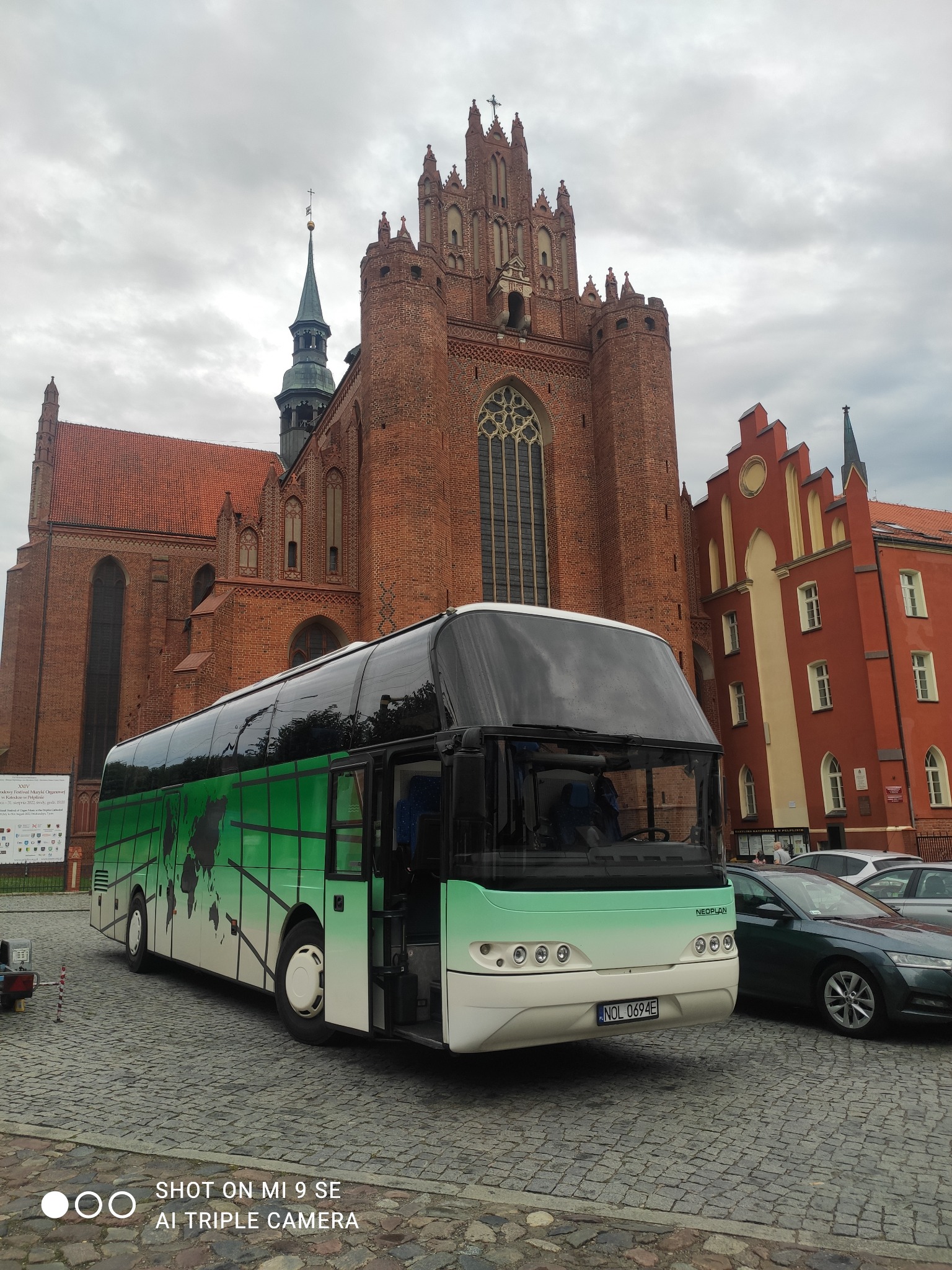 Autokar Neoplan z zielonym motywem mapy świata parkuje na brukowanym placu przed ceglaną gotycką katedrą z wieżą i spiczastą iglicą w pochmurny dzień.