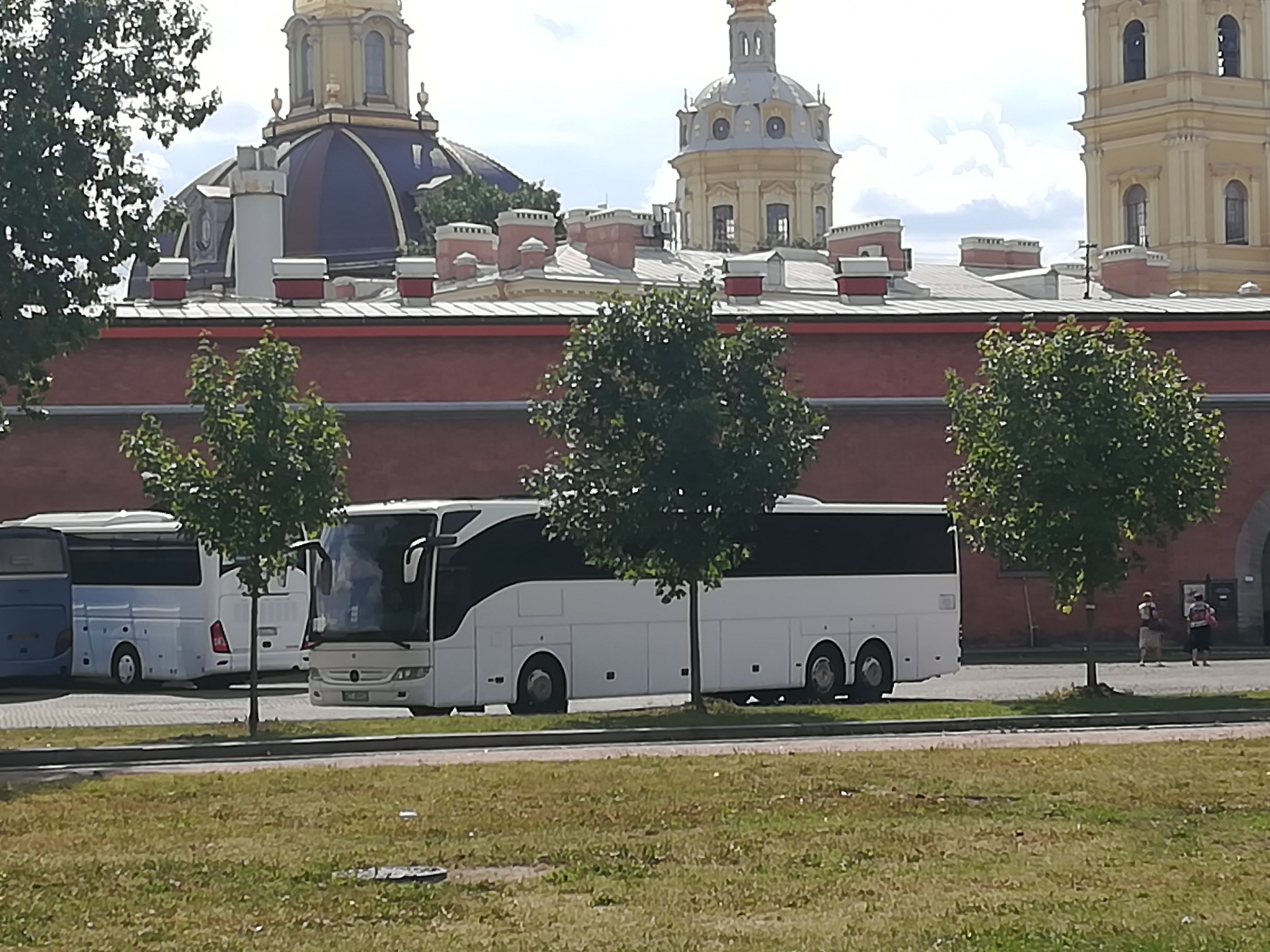 Biały autokar turystyczny Mercedes-Benz z czarnymi szybami, zaparkowany przed czerwonym murem z widocznymi wieżami Soboru Pietropawłowskiego w tle.