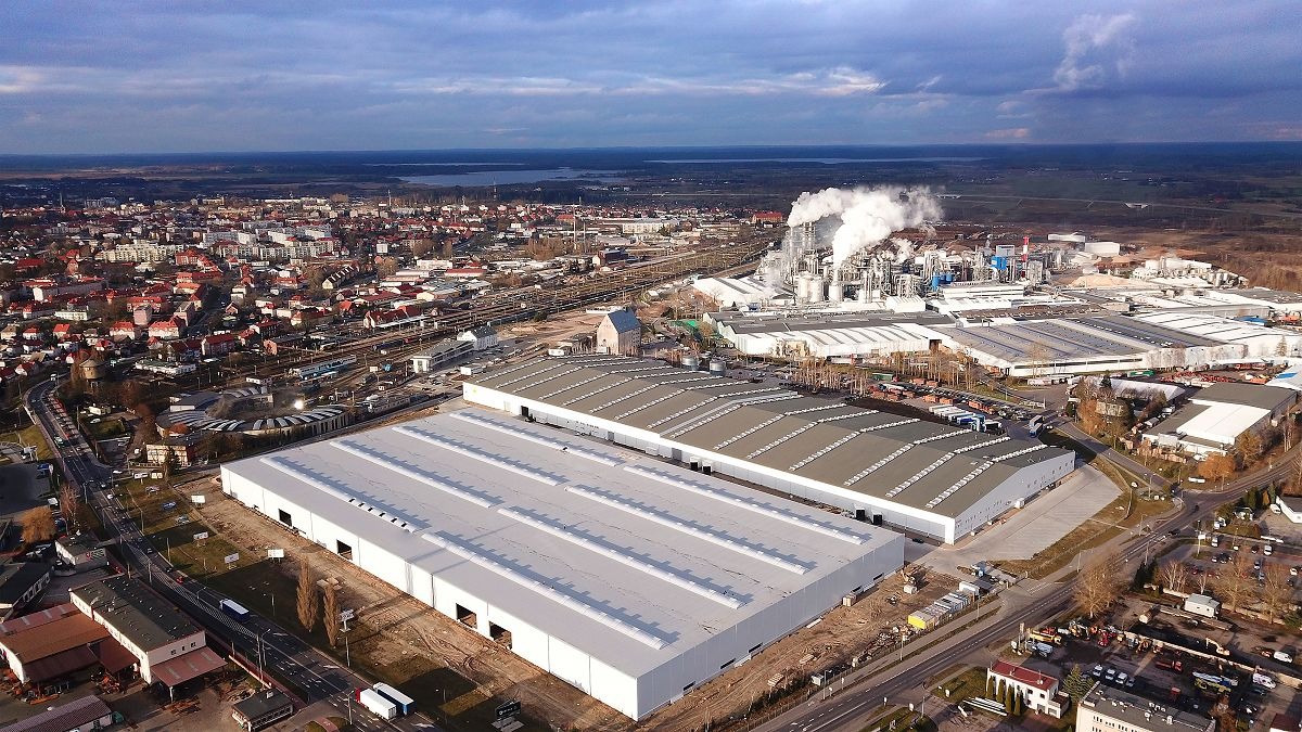 Panoramiczny widok z lotu ptaka na kompleks budynków przemysłowych z dachami z tarcicy budowlanej, z miastem i fabryką z dymiącymi kominami w tle.