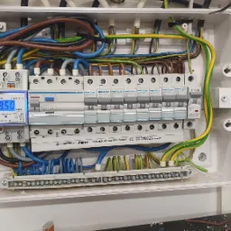 Otwarta skrzynka rozdzielcza z widocznymi bezpiecznikami Hager, miernikiem zużycia energii Orno, oraz okablowaniem w kolorach niebieskim, brązowym, żółto-zielonym i szarym, zamontowana na ścianie.