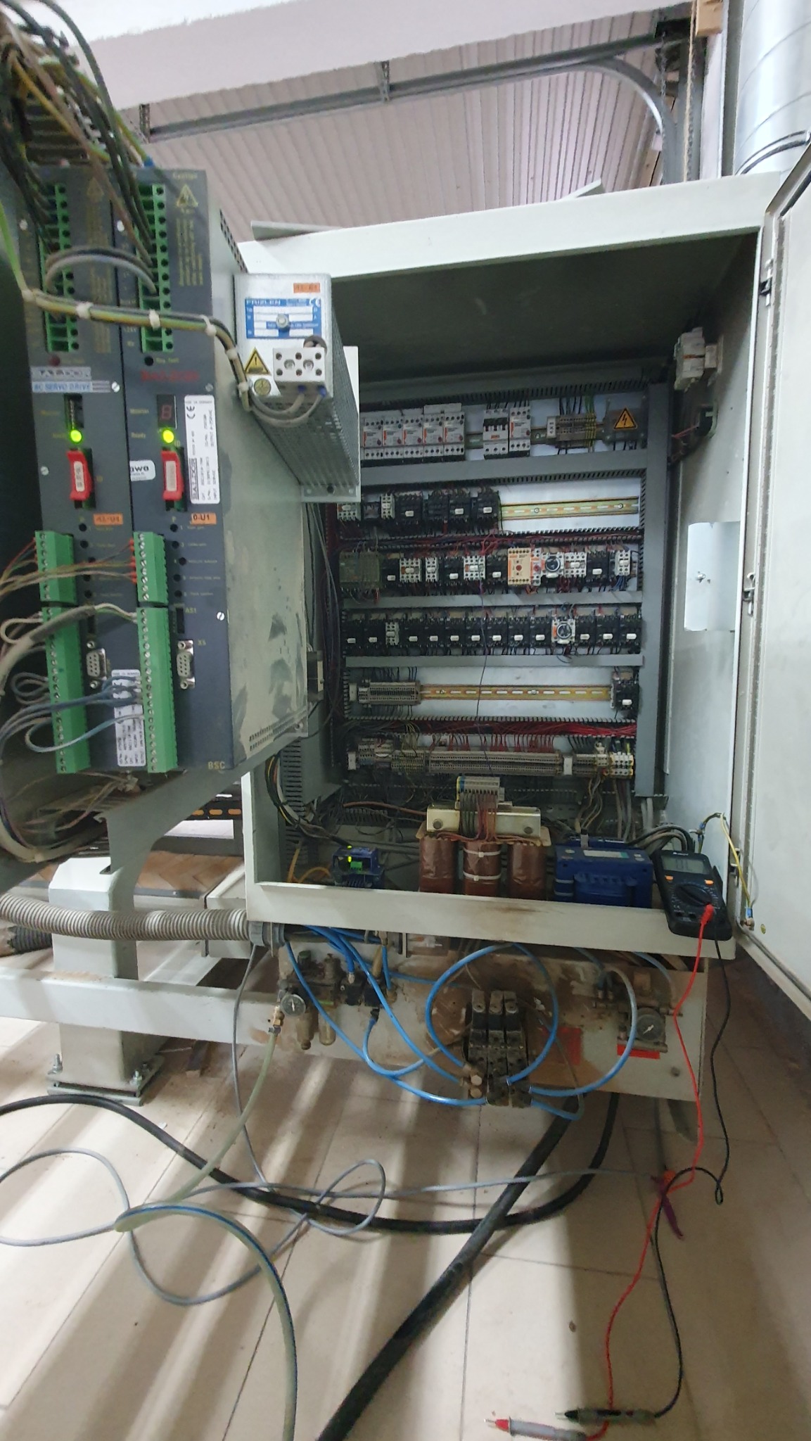 Otwarta szafa sterownicza maszyny przemysłowej z widocznymi elementami elektrycznymi, przewodami, transformatorami i multimetrem pomiarowym w prawym dolnym rogu.