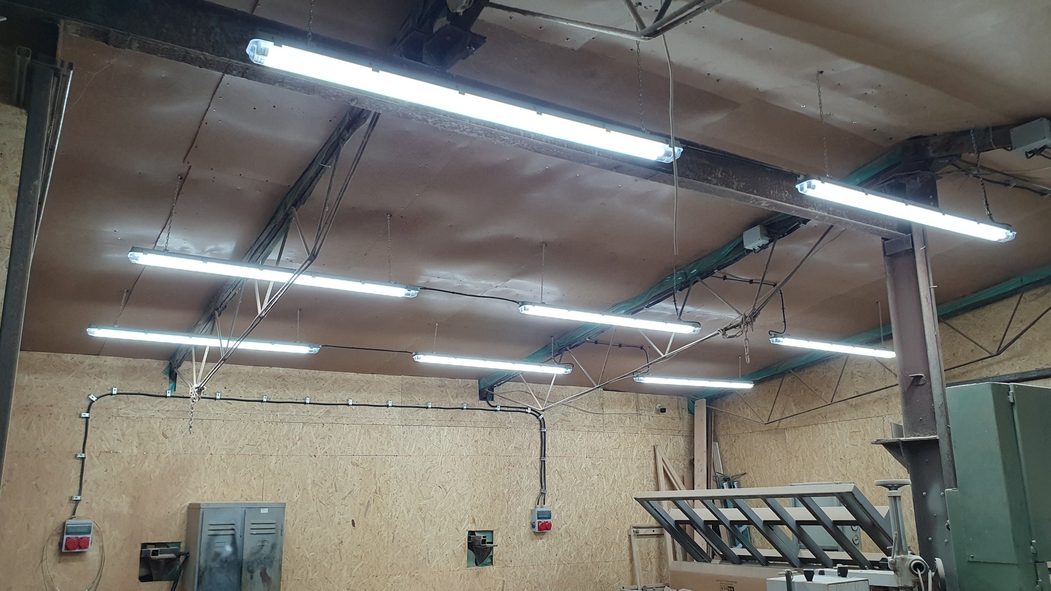 Wnętrze hali produkcyjnej z widocznymi lampami jarzeniowymi zamontowanymi pod sufitem, instalacją elektryczną poprowadzoną wzdłuż ściany z płyty OSB oraz maszyną stolarską w tle.