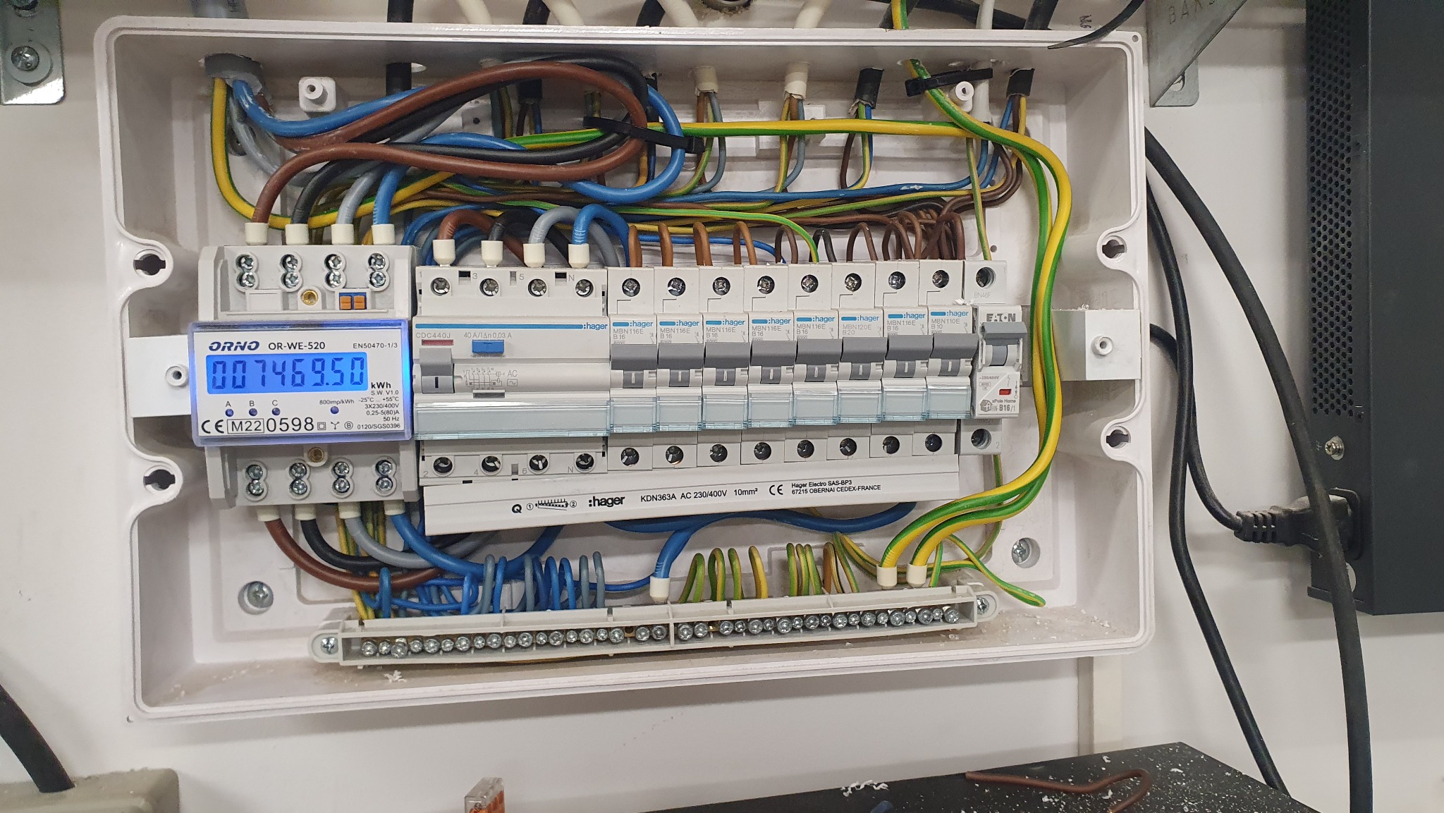 Otwarta skrzynka rozdzielcza z widocznymi bezpiecznikami Hager, miernikiem zużycia energii Orno, oraz okablowaniem w kolorach niebieskim, brązowym, żółto-zielonym i szarym, zamontowana na ścianie.