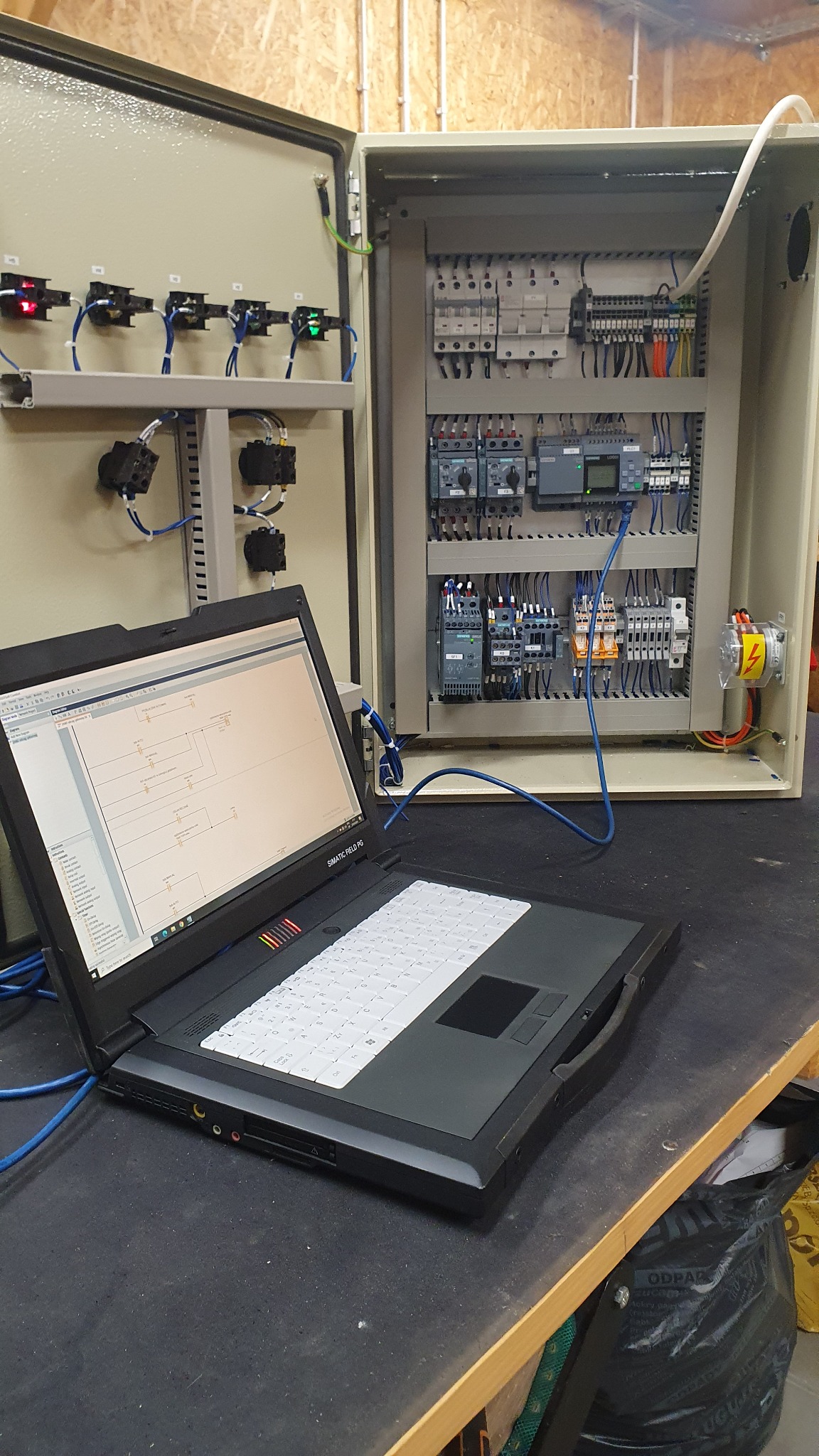 Otwarta szafa sterownicza z modułami Siemens, podłączona kablem ethernet do laptopa Simatic Field PG z widocznym schematem na ekranie.