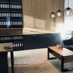 Nowoczesne biuro z regałami wypełnionymi segregatorami, drewnianymi szafkami, trzema lampami wiszącymi w metalowych kloszach oraz stolikiem kawowym z dekoracjami, oświetlone naturalnym światłem.