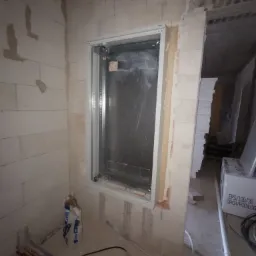 Wnęka w surowej ścianie z widoczną metalową obudową rozdzielnicy elektrycznej w trakcie montażu, z przewodami i workiem zaprawy na podłodze.