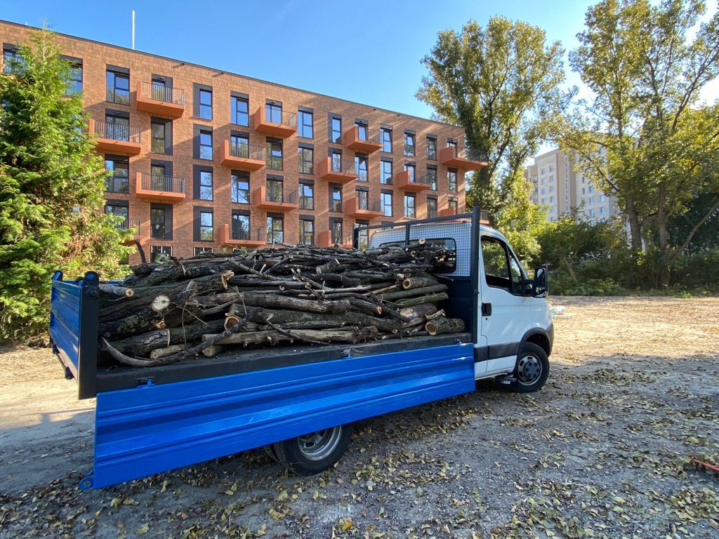 Niebieska ciężarówka z załadunkiem pociętych gałęzi na tle nowego budynku mieszkalnego i drzew.