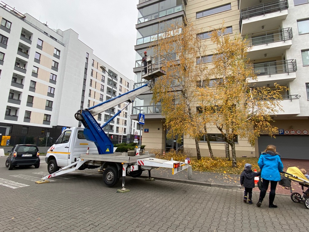 Pracownik na platformie podnośnika koszowego przycina gałęzie drzewa w pobliżu bloku mieszkalnego, z widocznymi ściętymi gałęziami na chodniku i przechodzącą obok rodziną.