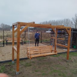 Osoba prywatna - Drewniana huśtawka ogrodowa z łańcuchami, na piaszczystym podłożu. W tle widoczna osoba i składowane drewno. Całość w naturalnej kolorystyce drewna.