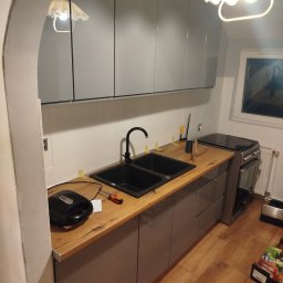 Osoba prywatna - Szare szafki kuchenne z drewnianym blatem i czarnym zlewem. Widoczny fragment okna i sprzęt AGD. Ujęcie z perspektywy podłogi, ukazujące detale wykończenia.