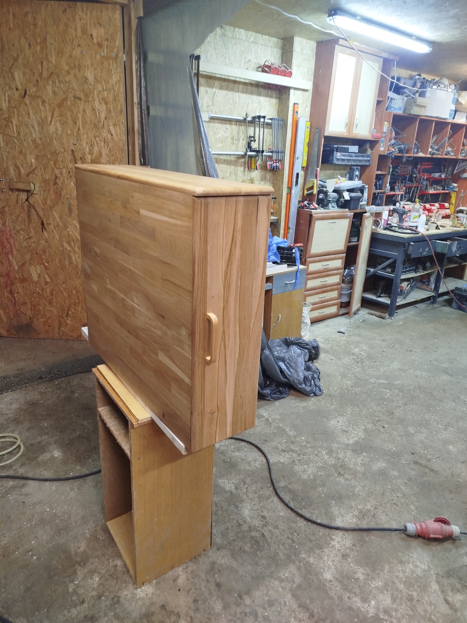 Drewniana szafka z uchwytem, w warsztacie stolarskim, na tle narzędzi i materiałów. Widoczny detal wykończenia i surowy charakter otoczenia.