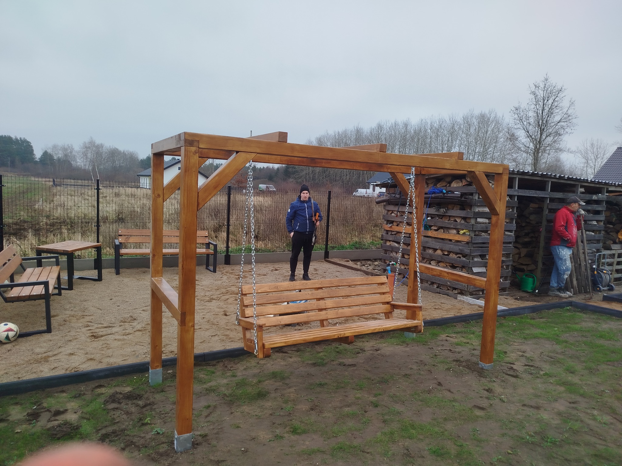 Drewniana huśtawka ogrodowa z łańcuchami, na piaszczystym podłożu. W tle widoczna osoba i składowane drewno. Całość w naturalnej kolorystyce drewna.