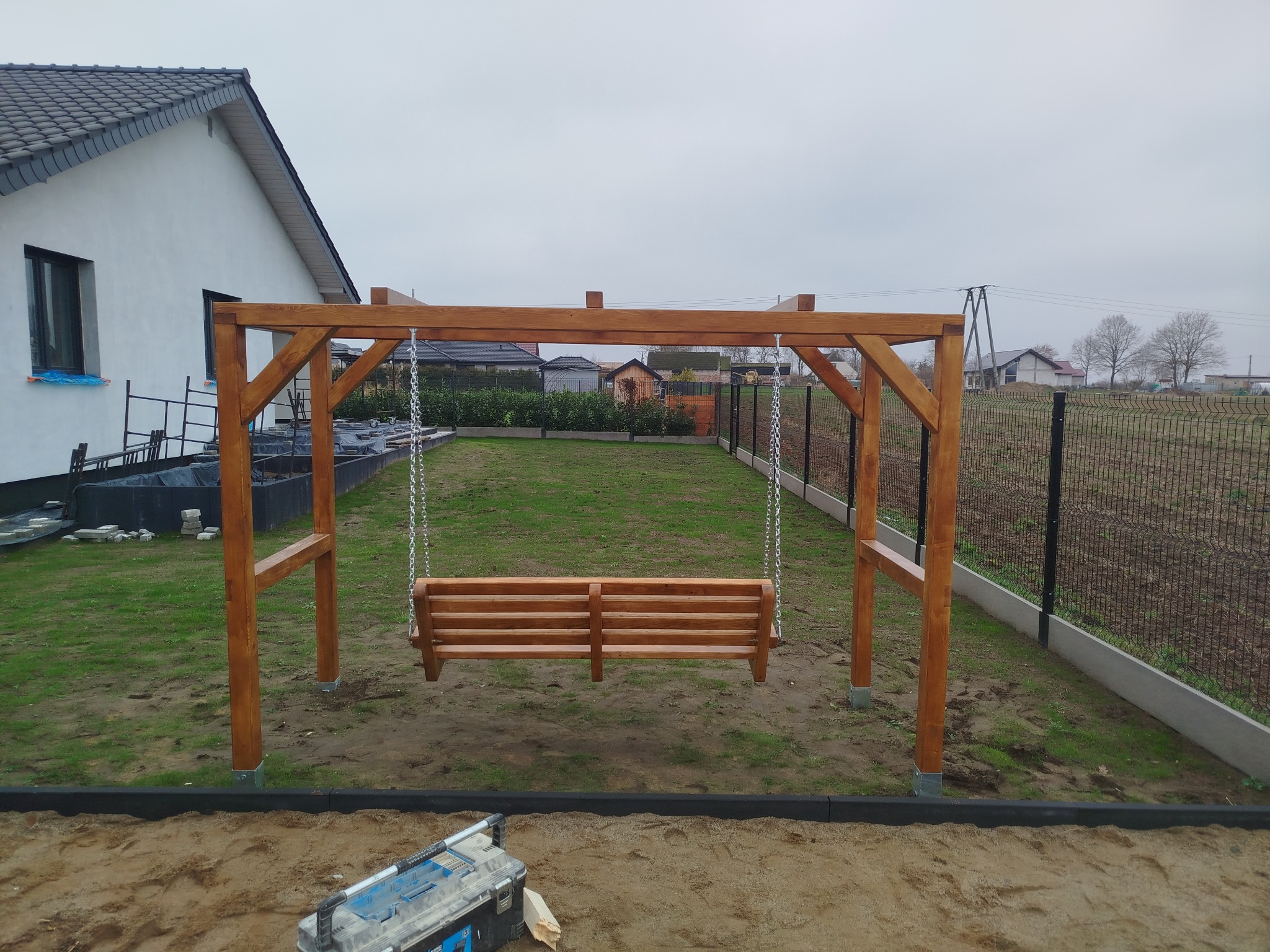 Drewniana huśtawka ogrodowa na łańcuchach, świeżo po montażu, z widoczną skrzynką narzędziową u podstawy, na tle trawnika i ogrodzenia.