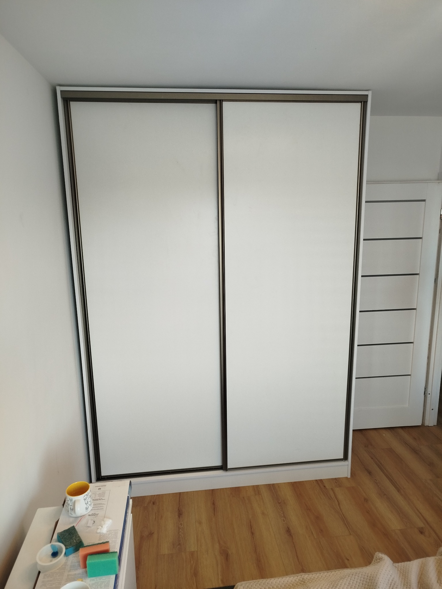 Biała szafa wnękowa z przesuwnymi drzwiami i ciemną ramą, widoczna z bliska na tle drewnianej podłogi i białej ściany. Obok szafy stolik z akcesoriami.