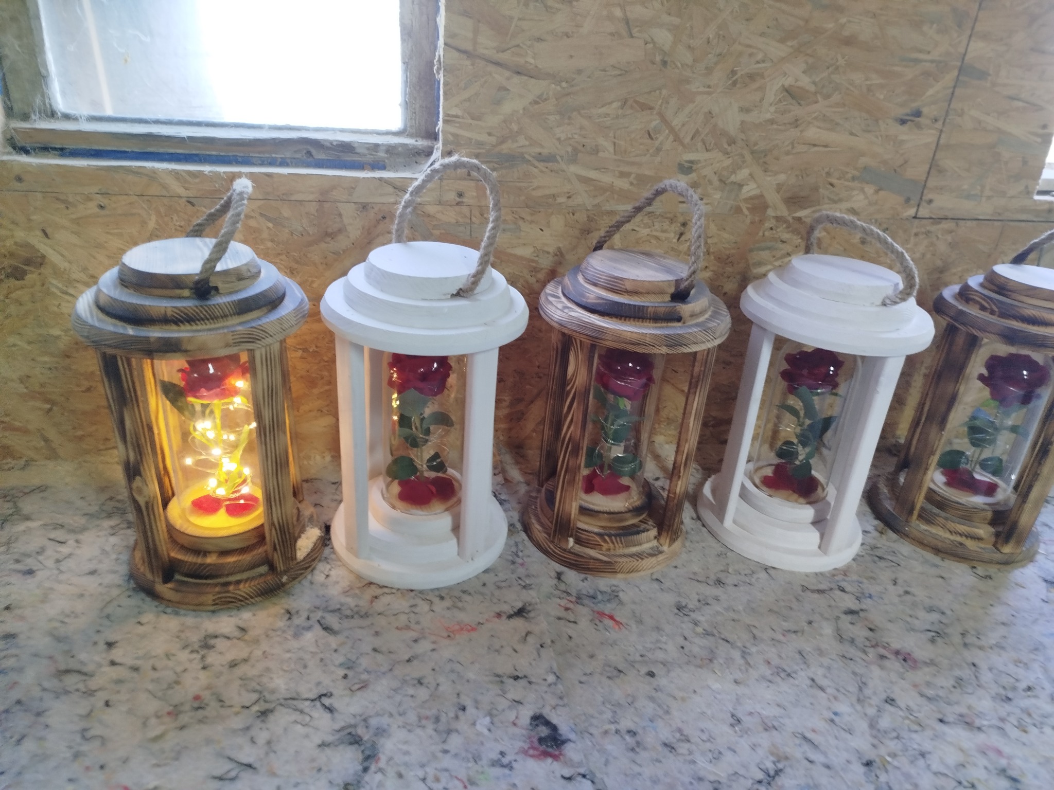 Rząd drewnianych lampionów z różami w szklanych tubach, niektóre z oświetleniem LED, na tle płyty OSB. Różne wykończenia drewna: naturalne i białe.