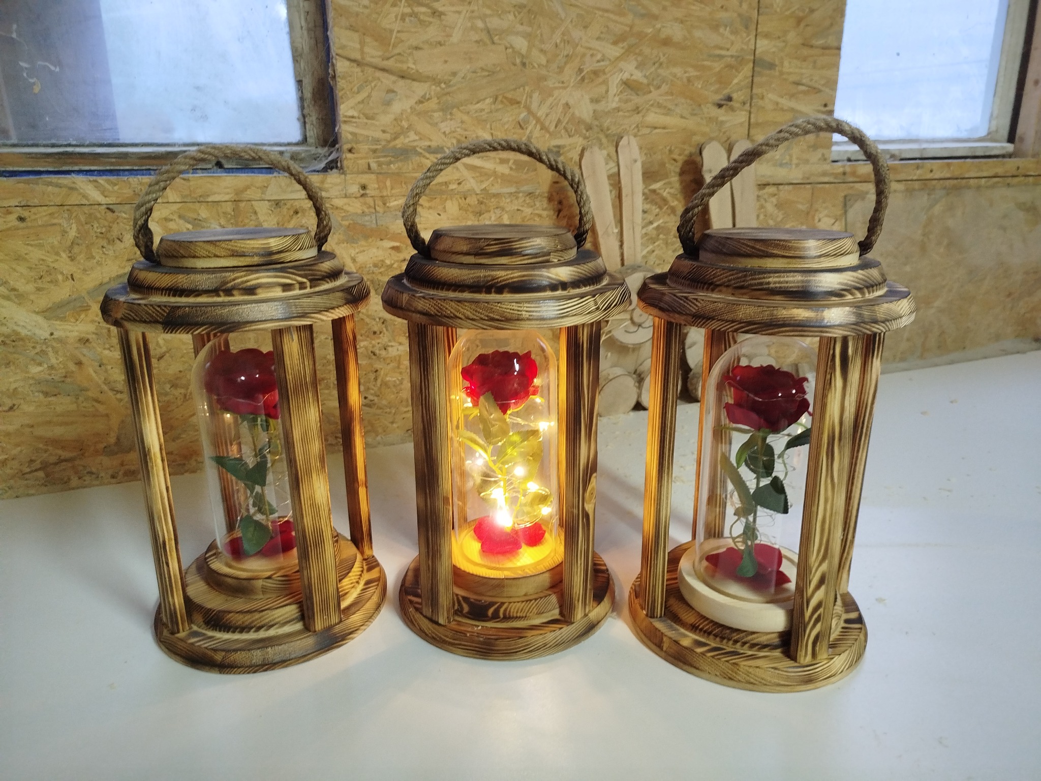 Trzy drewniane lampiony z różami pod szklanym kloszem, oświetlone ciepłym światłem, z uchwytami z grubego sznura, na tle drewnianej ściany i okna.