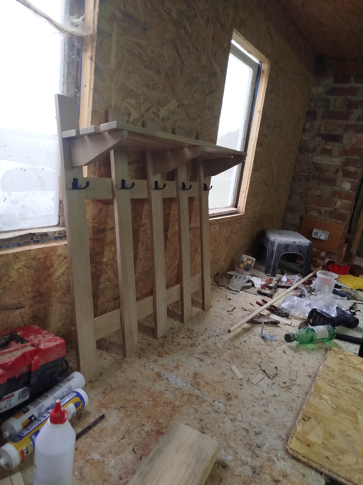 Drewniany wieszak z półką, surowy, z haczykami, ustawiony w warsztacie z oknami i narzędziami, w trakcie tworzenia, z widocznymi śladami obróbki drewna.