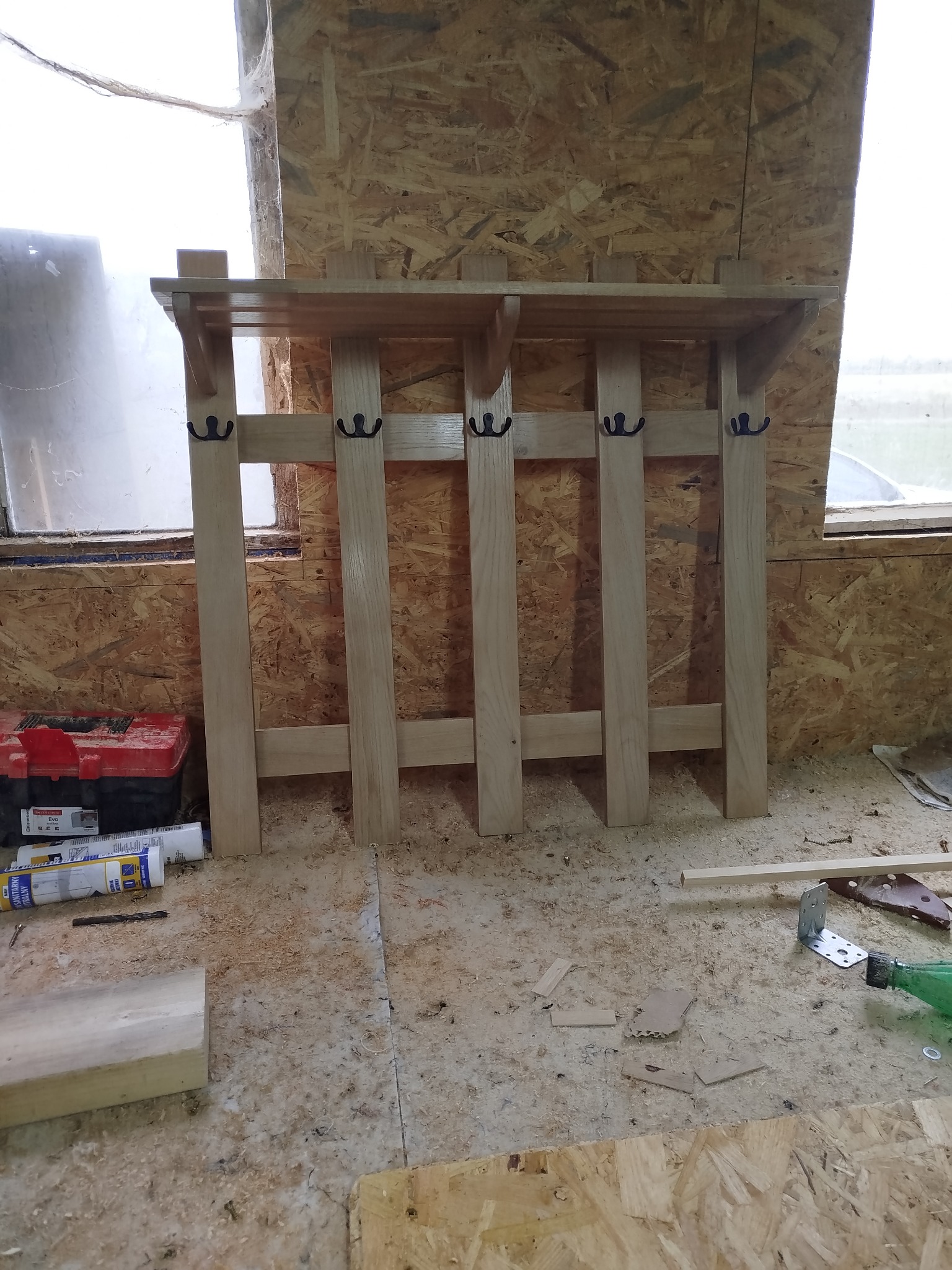 Surowy, drewniany wieszak z półką i czarnymi haczykami, stojący w warsztacie na tle płyty OSB i okna. Widoczne narzędzia i akcesoria stolarskie.