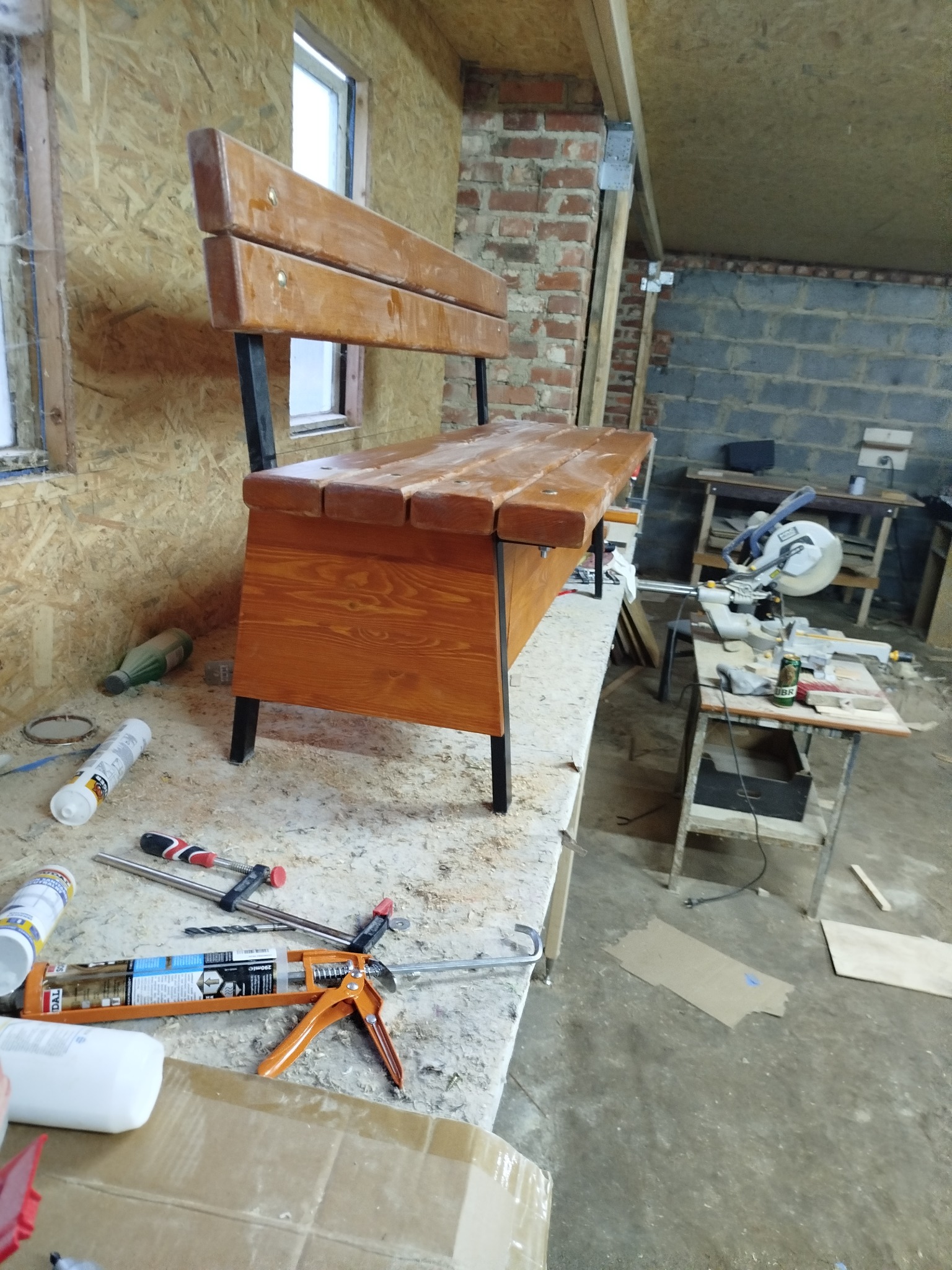 Drewniana ławka z oparciem na metalowych nogach, w warsztacie. Widoczne narzędzia stolarskie i materiały budowlane. Ściany z cegły i płyty OSB.