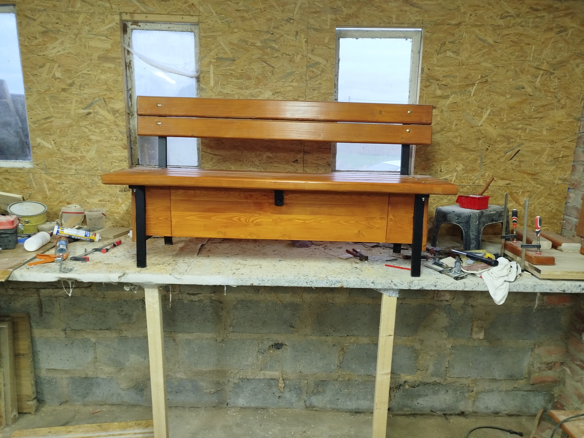 Drewniana ławka z oparciem i schowkiem, na czarnych metalowych nogach, w warsztacie. Widoczne narzędzia i okna w tle. Ciepłe oświetlenie podkreśla strukturę drewna.