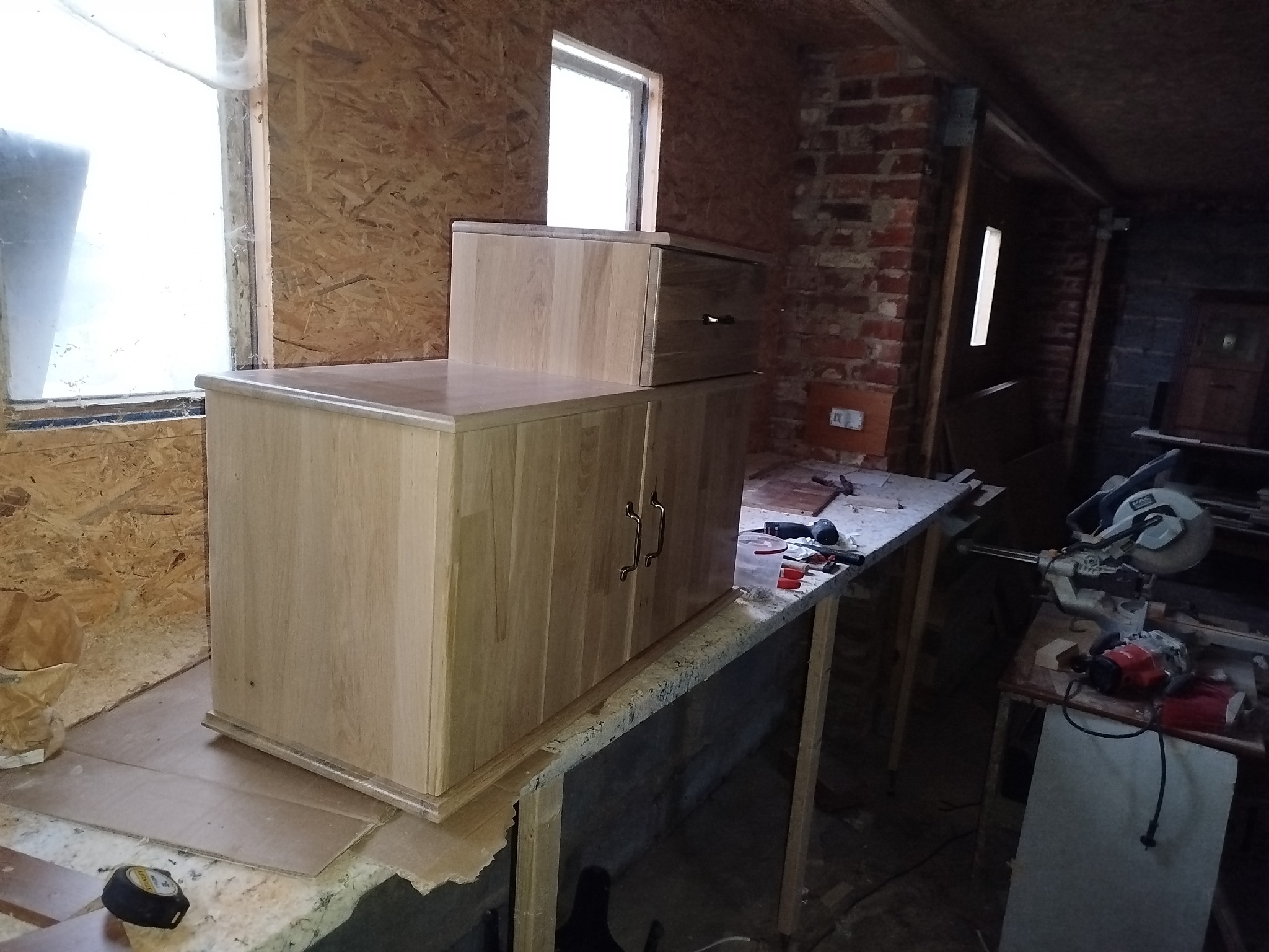 Drewniana komoda z szufladą w warsztacie, widoczne narzędzia stolarskie i okno. Surowe wnętrze z ceglaną ścianą w tle. Ujęcie z bliska, detale wykończenia.