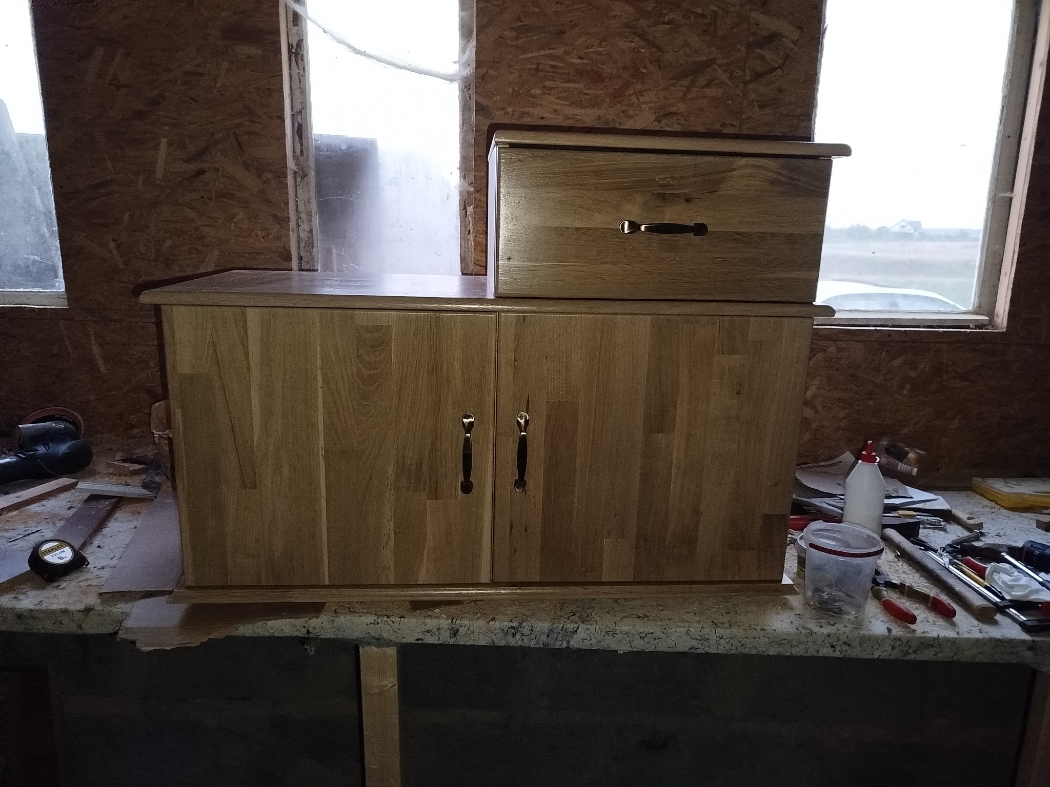 Drewniana szafka z szufladą na blacie roboczym, widoczne narzędzia i okno w tle. Wykończenie w naturalnym odcieniu drewna, proste uchwyty.