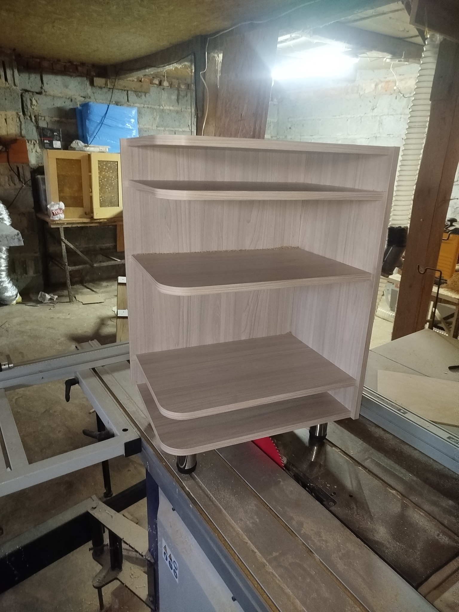 Półka z zaokrąglonymi rogami, wykonana z jasnego drewna, stoi na pile stołowej w warsztacie. Widoczne surowe ściany i elementy wyposażenia stolarskiego.