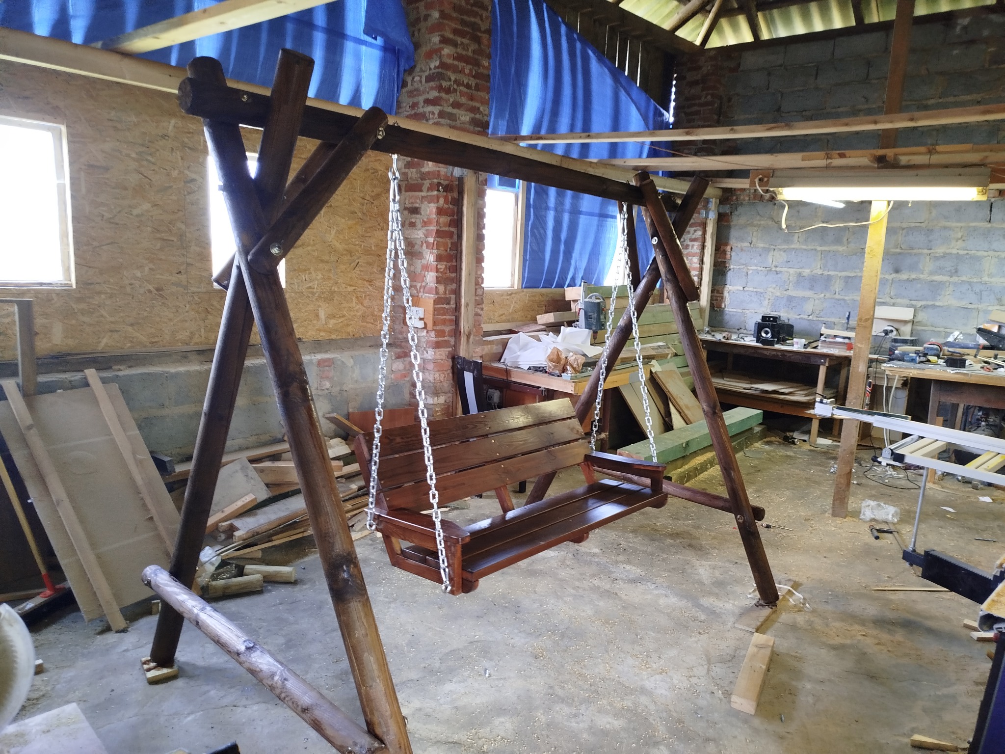Drewniana huśtawka ogrodowa z oparciem, zawieszona na łańcuchach, w warsztacie. Widoczne narzędzia i materiały stolarskie w tle. Wykończenie w ciemnym brązie.