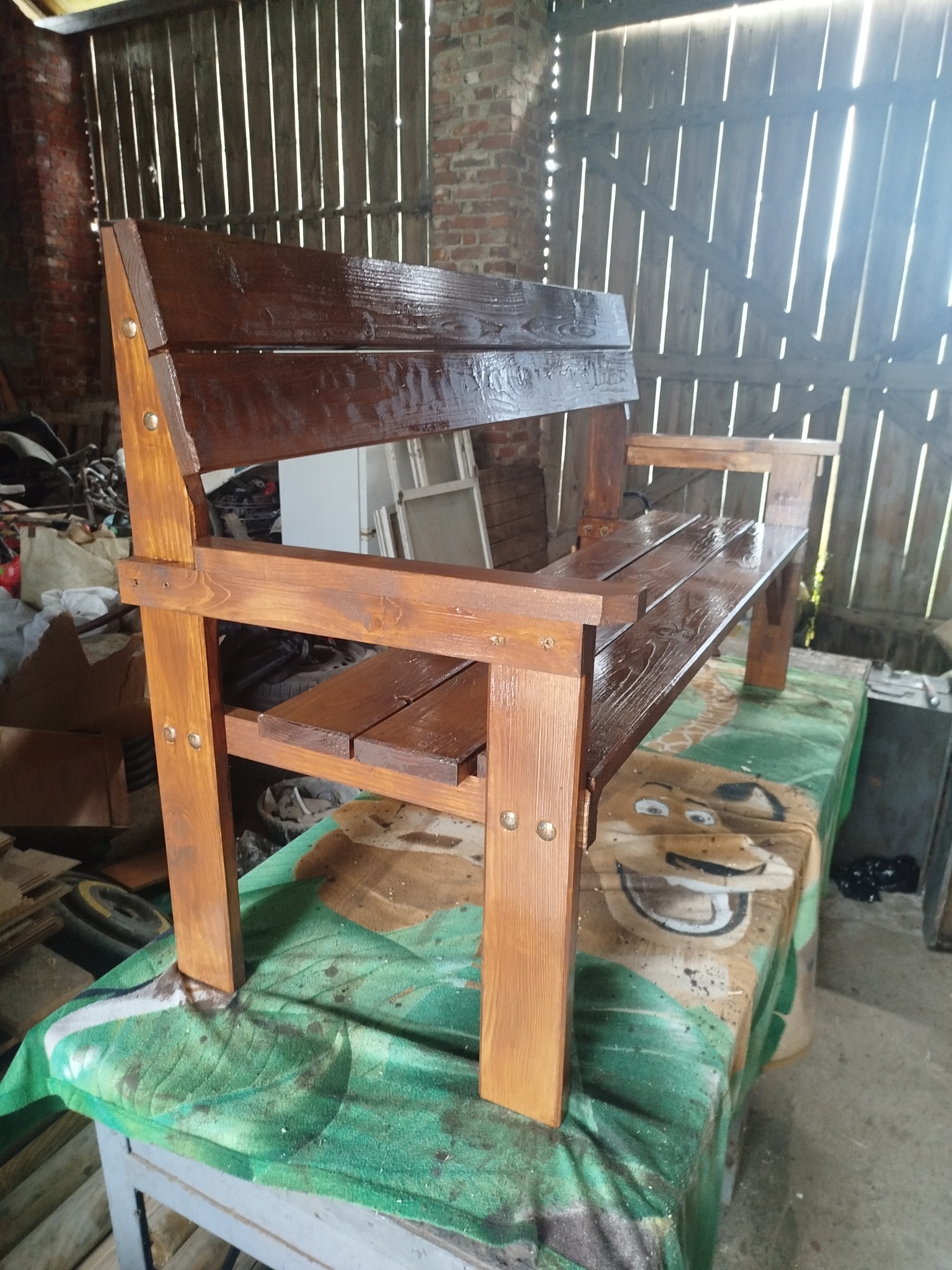 Drewniana ławka ogrodowa z oparciem, świeżo polakierowana na brązowo, z widocznymi śrubami, ustawiona w warsztacie na stole przykrytym zielonym materiałem.