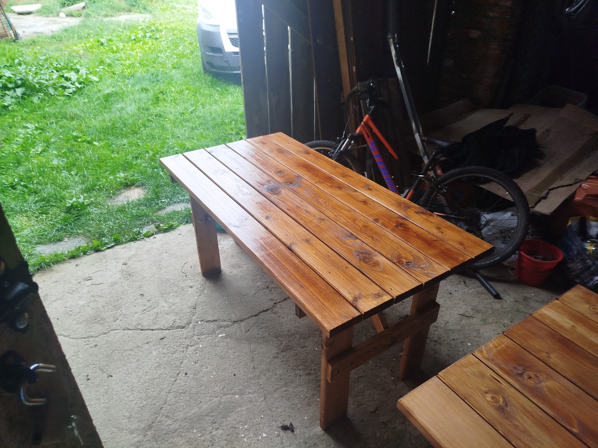 Drewniany stół z blatem z desek, świeżo polakierowany na brązowo, stoi na betonowej posadzce, obok trawnik i rower oparty o ścianę. Widoczne słoje drewna.