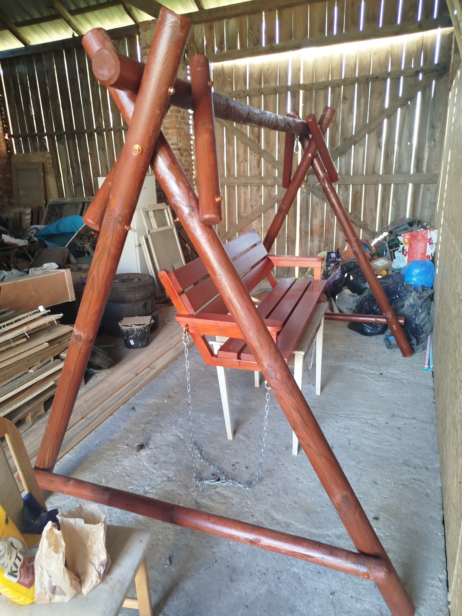 Drewniana huśtawka ogrodowa, wykonana z okrąglaków, pomalowana na brązowo, zawieszona na łańcuchach. W tle widoczne deski i inne przedmioty.