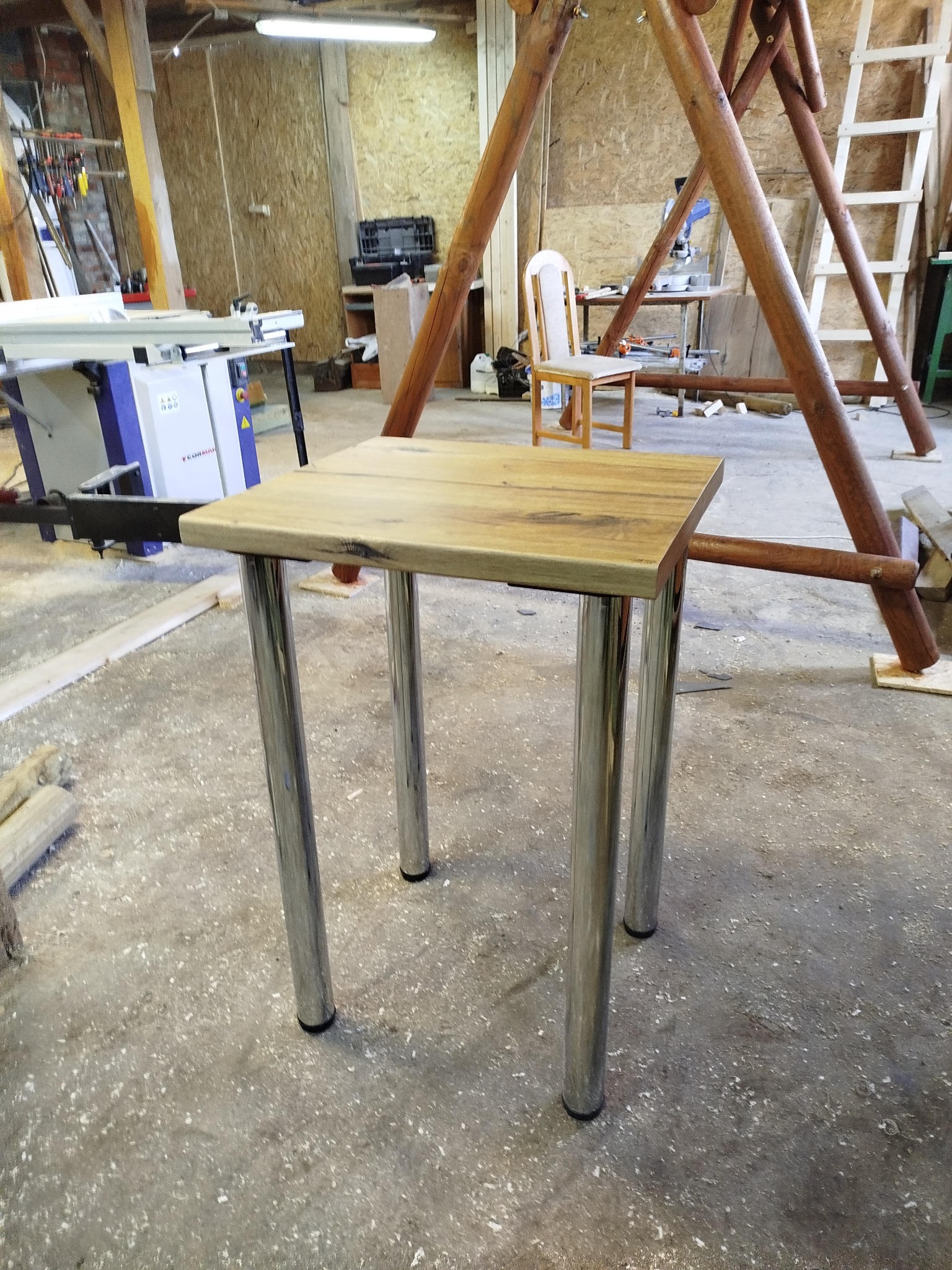 Drewniany stolik kawowy z metalowymi nogami na tle warsztatu stolarskiego z widoczną piłą stołową i elementami konstrukcji drewnianej.