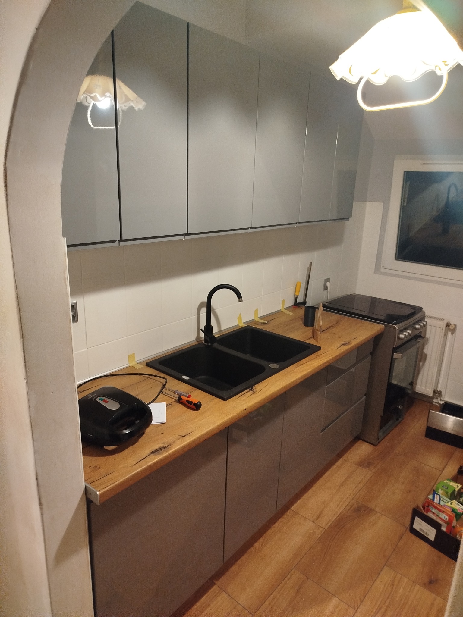 Szare szafki kuchenne z drewnianym blatem i czarnym zlewem. Widoczny fragment okna i sprzęt AGD. Ujęcie z perspektywy podłogi, ukazujące detale wykończenia.