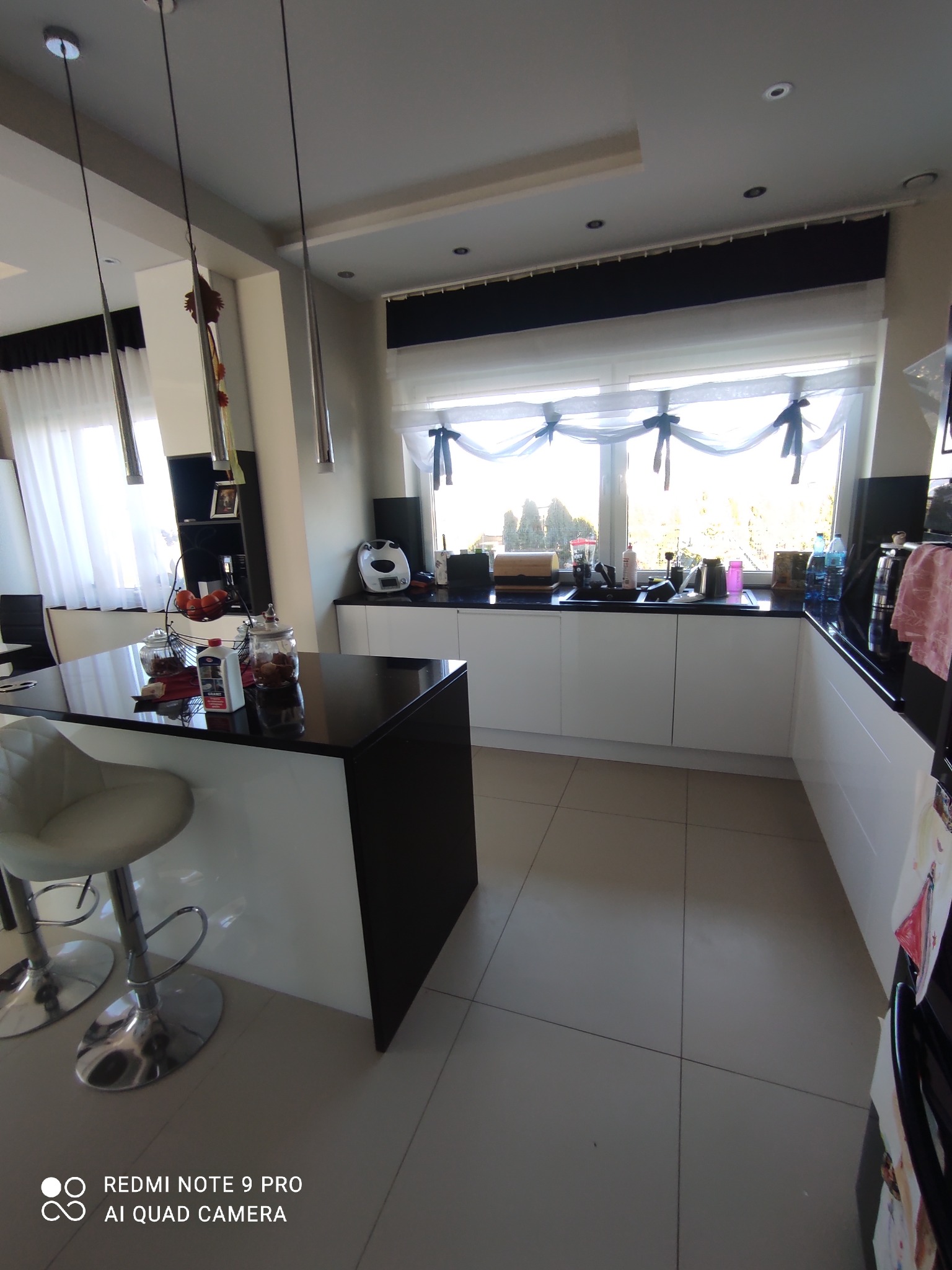 Jasna kuchnia z czarnym blatem, białymi szafkami i wyspą, okno z firankami z kokardami, widoczne owoce w koszyku i słoiki na blacie, płytki na podłodze.