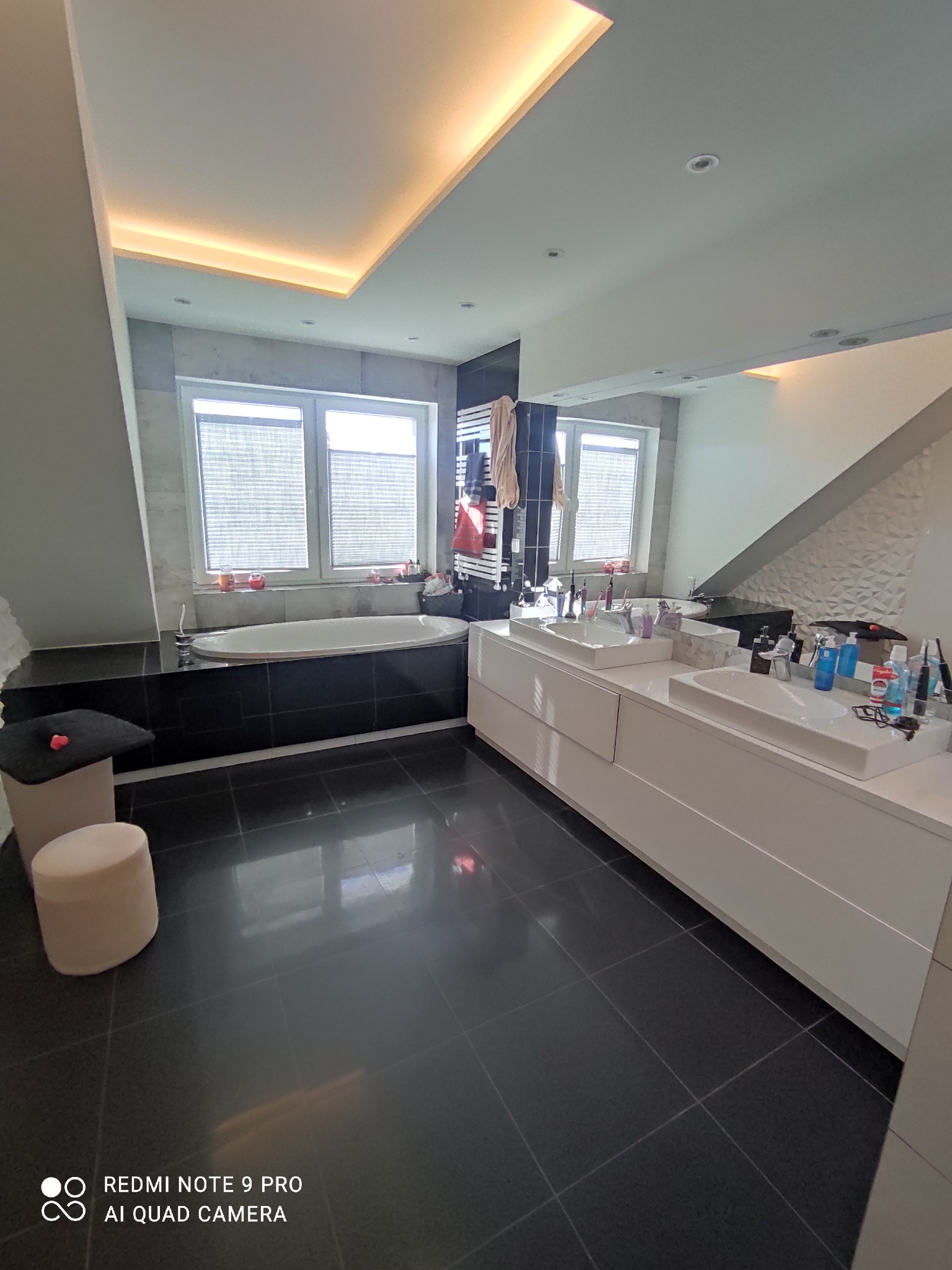 Nowoczesna łazienka z czarną podłogą, białą wanną i umywalkami, widoczne akcesoria kosmetyczne i ręczniki na grzejniku.