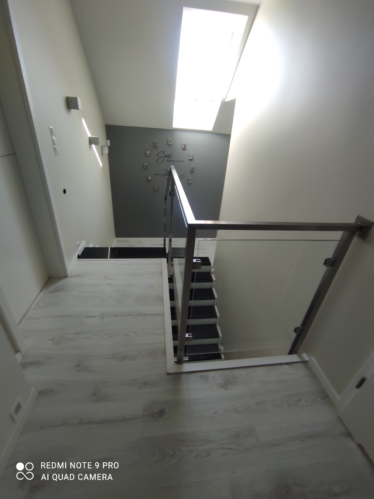 Wnętrze domu z widokiem na schody z czarnymi stopniami i szklaną balustradą ze srebrnymi elementami, oświetlone przez okno dachowe, ściana z dekoracyjnym napisem i ramkami na zdjęcia.