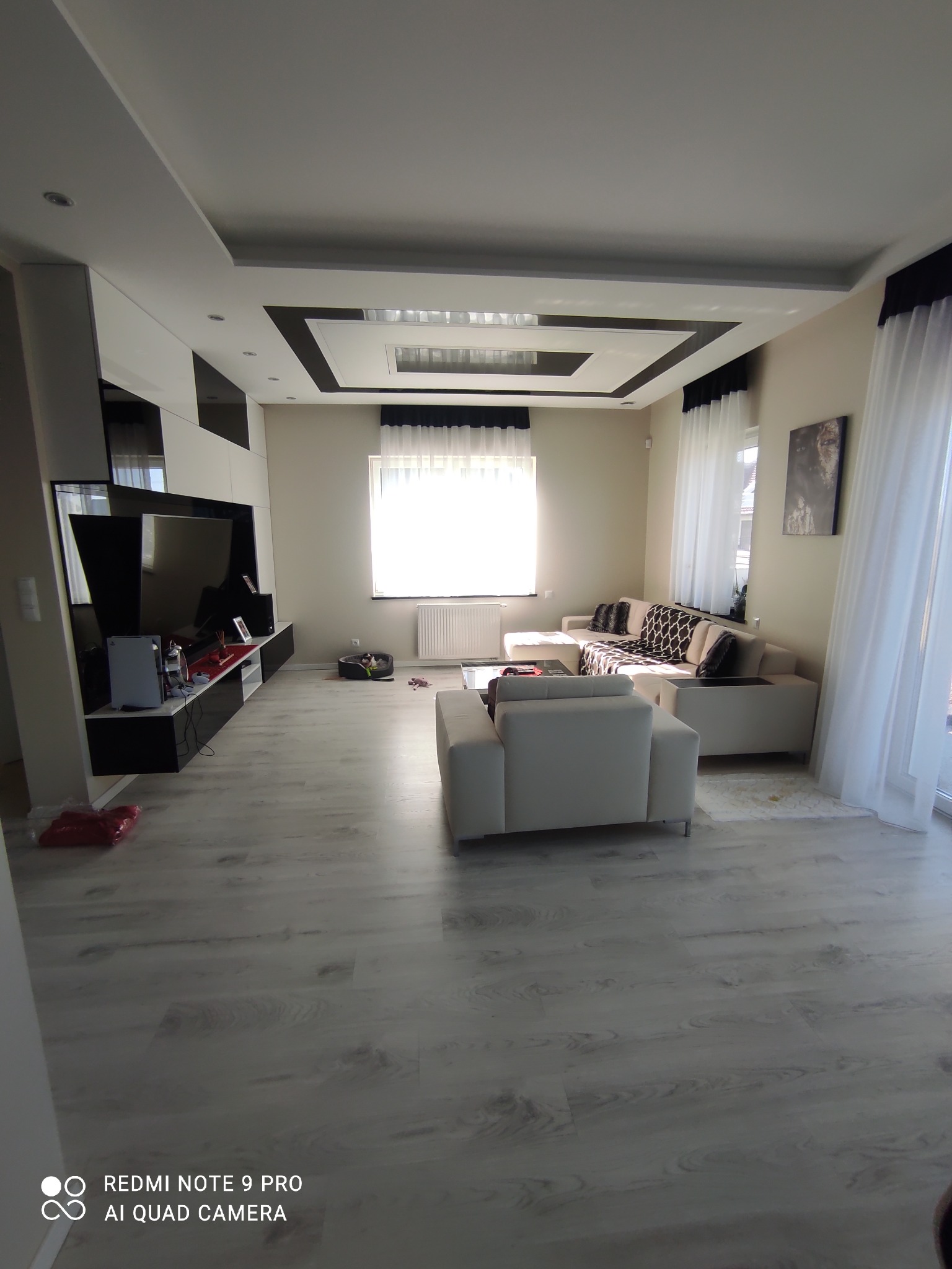 Jasne, przestronne wnętrze salonu z szarymi panelami podłogowymi, białymi meblami wypoczynkowymi i nowoczesną zabudową ściany z telewizorem; widoczny odkurzacz automatyczny oraz legowisko dla psa...