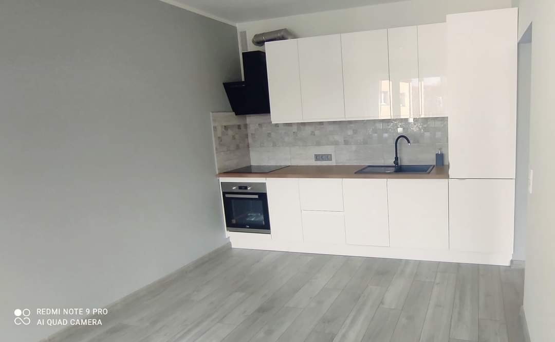 Nowoczesna, biała kuchnia z czarnymi akcentami: okap, bateria i piekarnik. Ściany szare, podłoga jasna, drewniana. Minimalistyczny design, brak dekoracji.