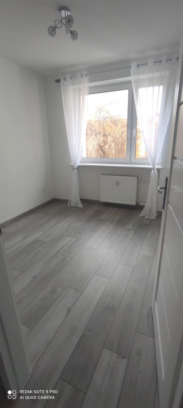 Puste, świeżo wykończone wnętrze pokoju z szarymi panelami podłogowymi, białymi ścianami, oknem z białymi firankami i grzejnikiem pod oknem, widok z perspektywy drzwi.