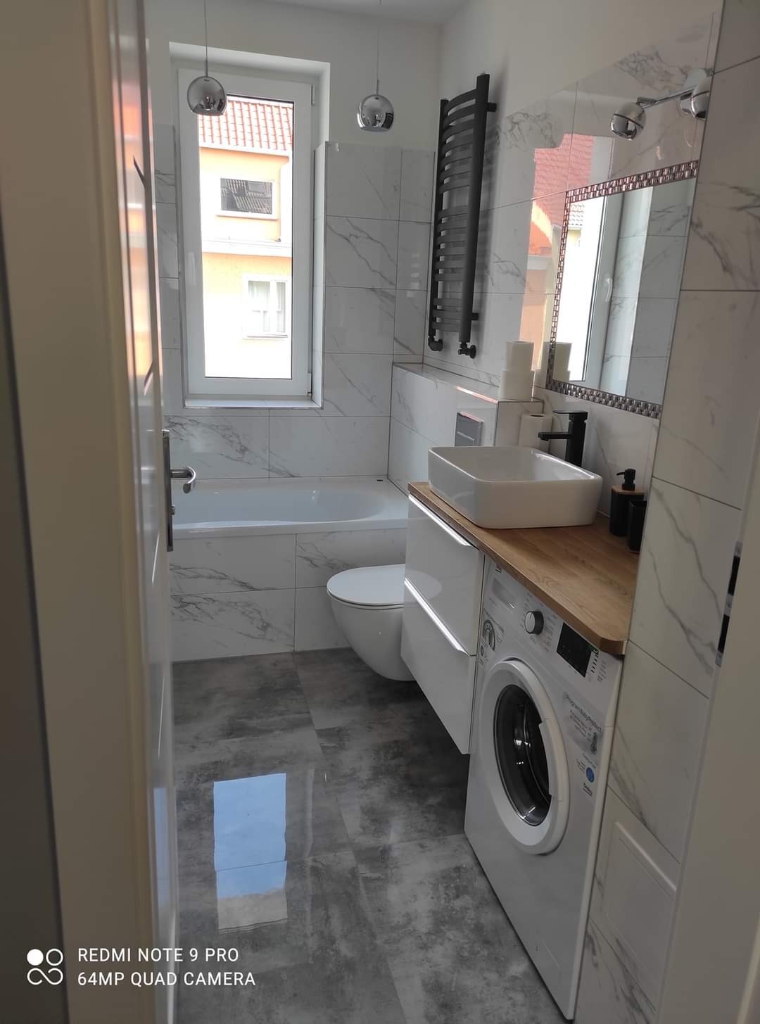 Nowoczesna łazienka z marmurowymi płytkami, białą armaturą, drewnianym blatem nad pralką i grzejnikiem drabinkowym, widziana z perspektywy wejścia.