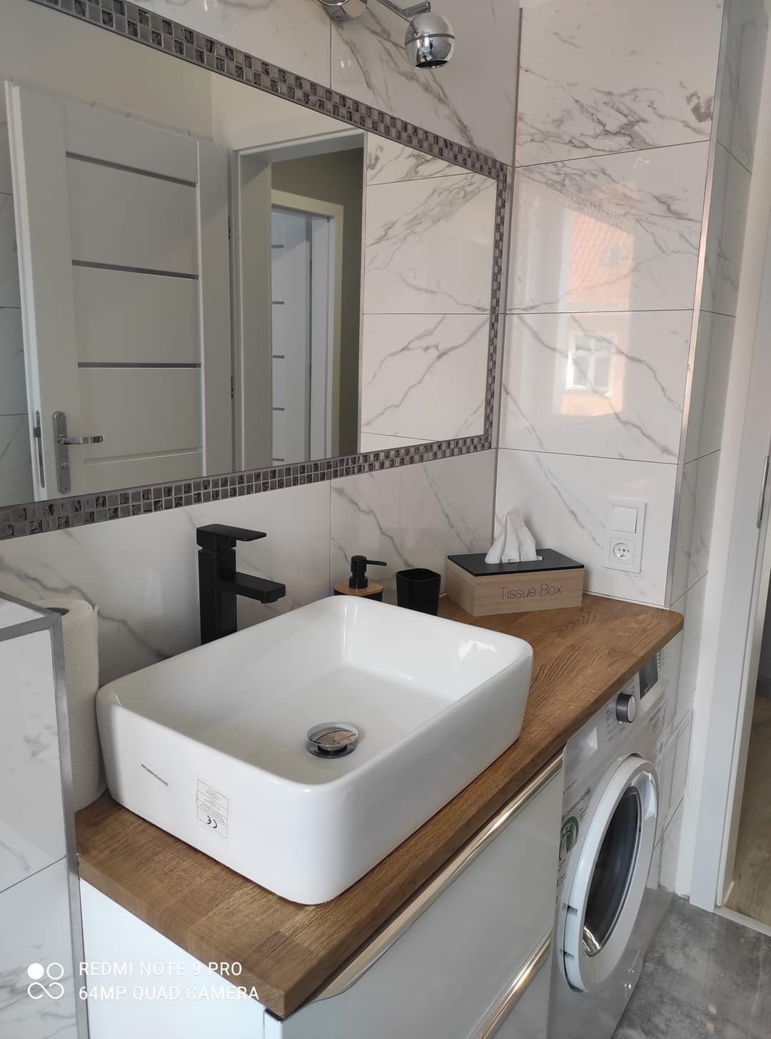 Biała, prostokątna umywalka nablatowa z czarną baterią, marmurowe płytki na ścianie, lustro z mozaikową ramą i drewniany blat z dozownikiem mydła, kubkiem i pudełkiem na chusteczki w czystej łazience.