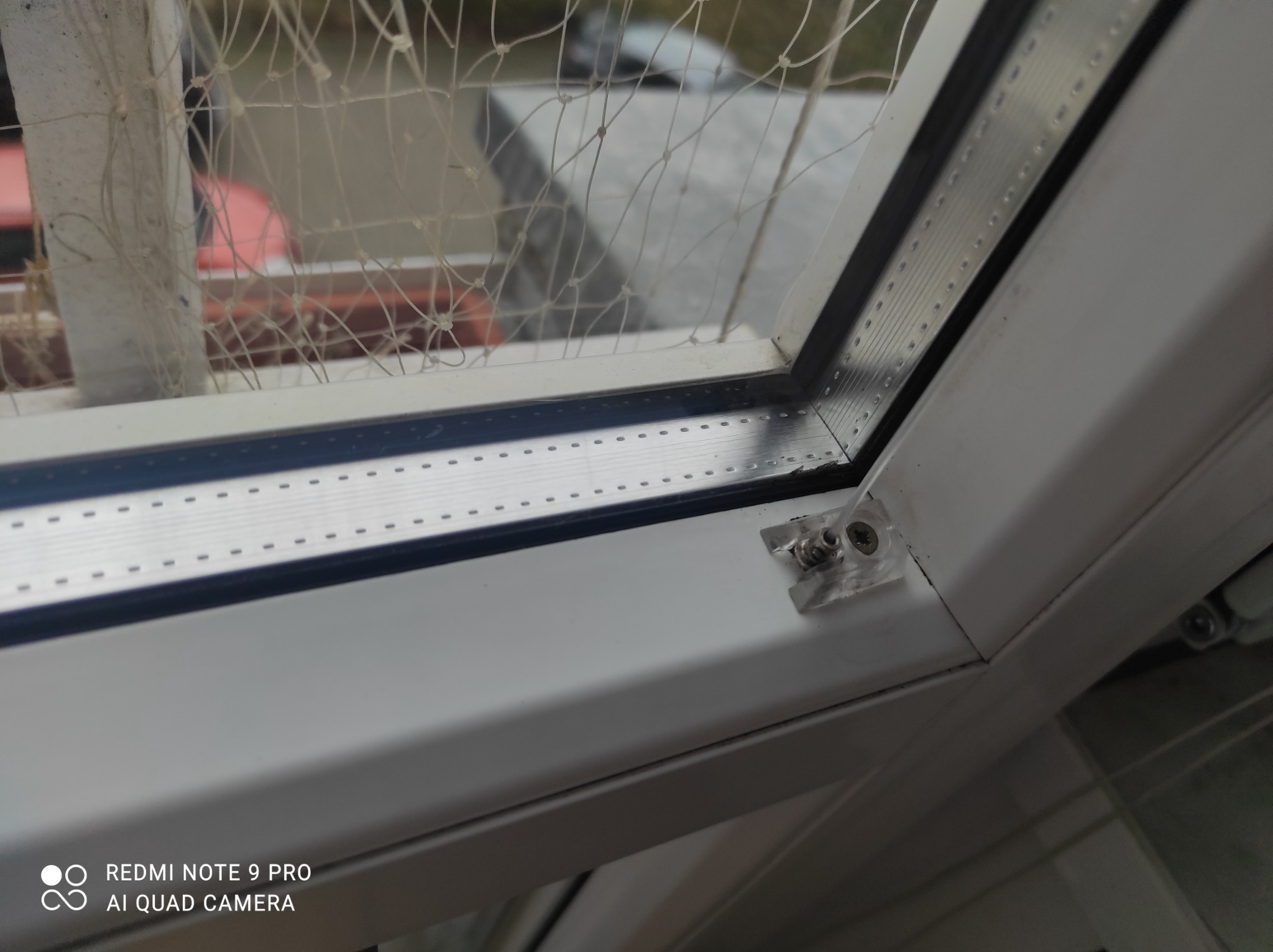 Widok z bliska na białą ramę okienną z zamontowaną siatką ochronną przed ptakami, widoczne elementy mocujące i perforowana listwa aluminiowa.