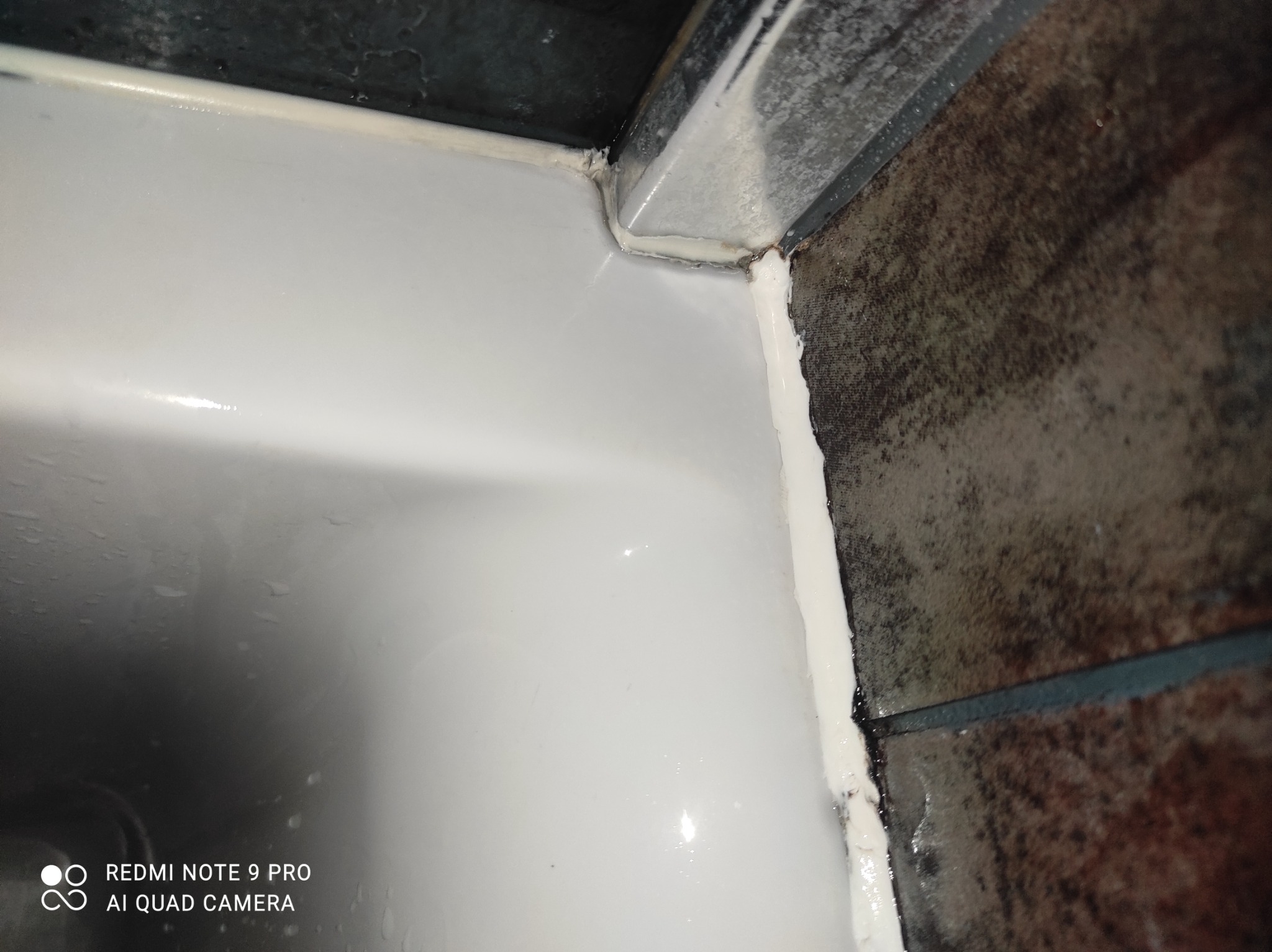 Szczegółowe ujęcie świeżo nałożonego, białego silikonu w rogu kabiny prysznicowej, widoczne ciemne płytki i fragment odpływu, wykonane z bliskiej odległości.