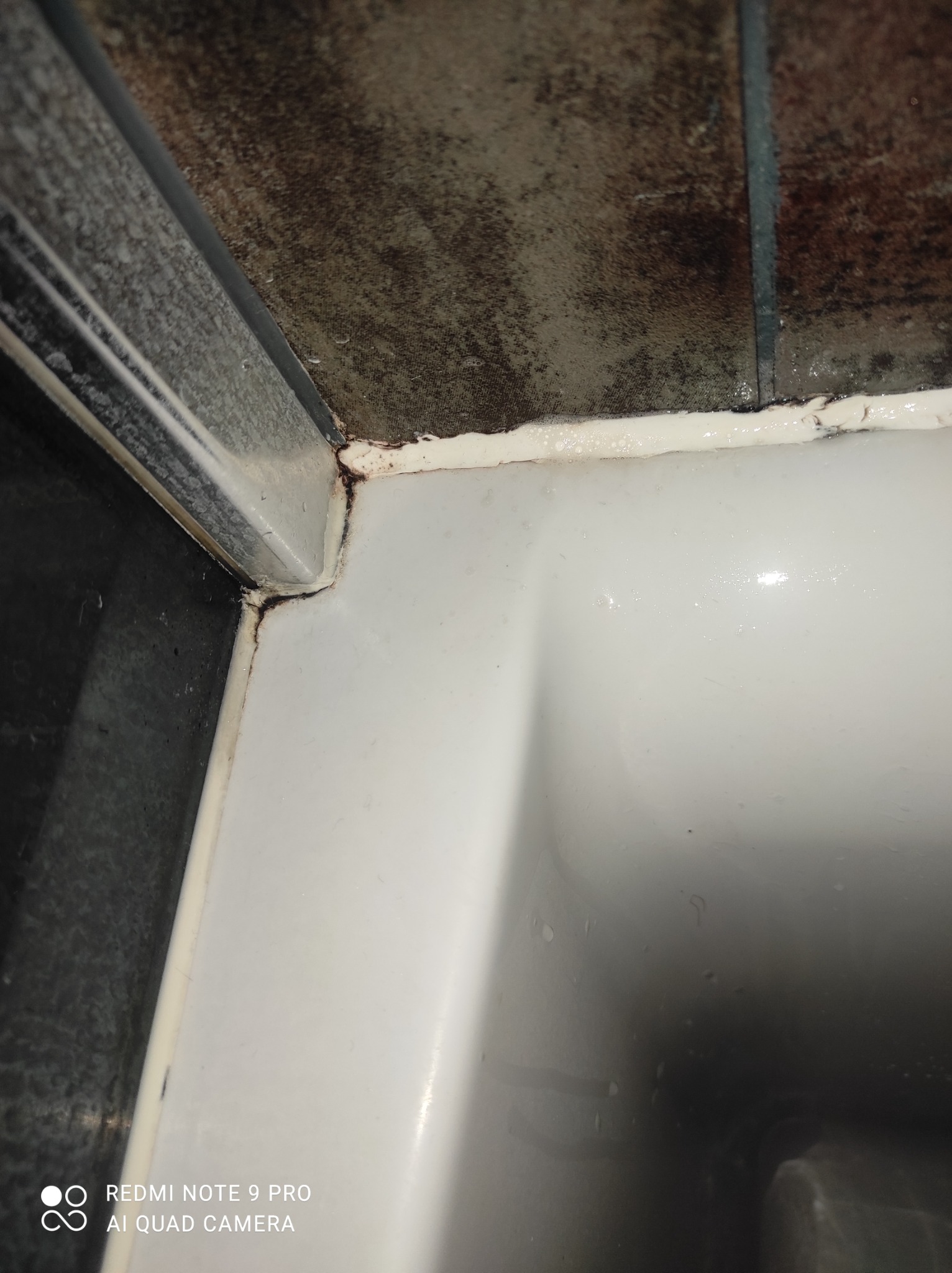 Szczegółowe ujęcie narożnika w łazience z widocznym zagrzybieniem i pleśnią na silikonie przy wannie i płytkach, wymagające interwencji i usunięcia.
