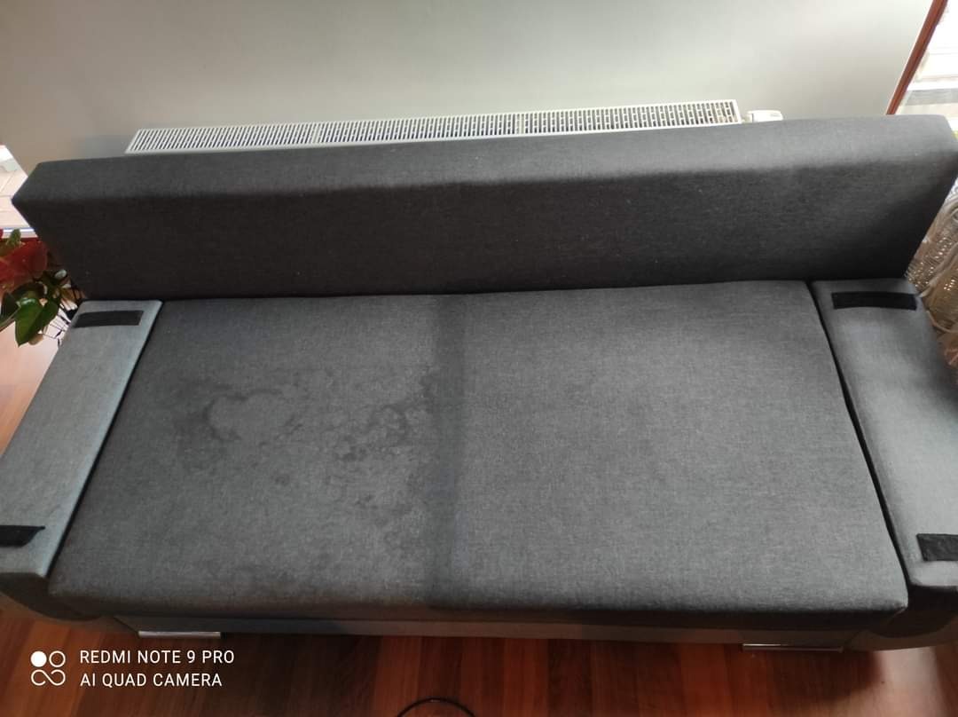 Szara rozkładana kanapa z widocznymi plamami na siedzisku, sfotografowana z góry w słabym oświetleniu, z widocznym grzejnikiem w tle.