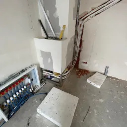 Instalacja rozdzielacza ogrzewania podłogowego w surowym stanie, widoczne rury, styropian izolacyjny, elementy konstrukcji ściany i ślady po kuciu w ścianie.
