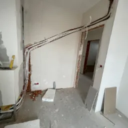 Wnętrze w trakcie remontu z widocznymi przewodami instalacji elektrycznej ułożonymi wzdłuż ścian i nad ościeżnicą, fragmenty płyt kartonowo-gipsowych na podłodze.